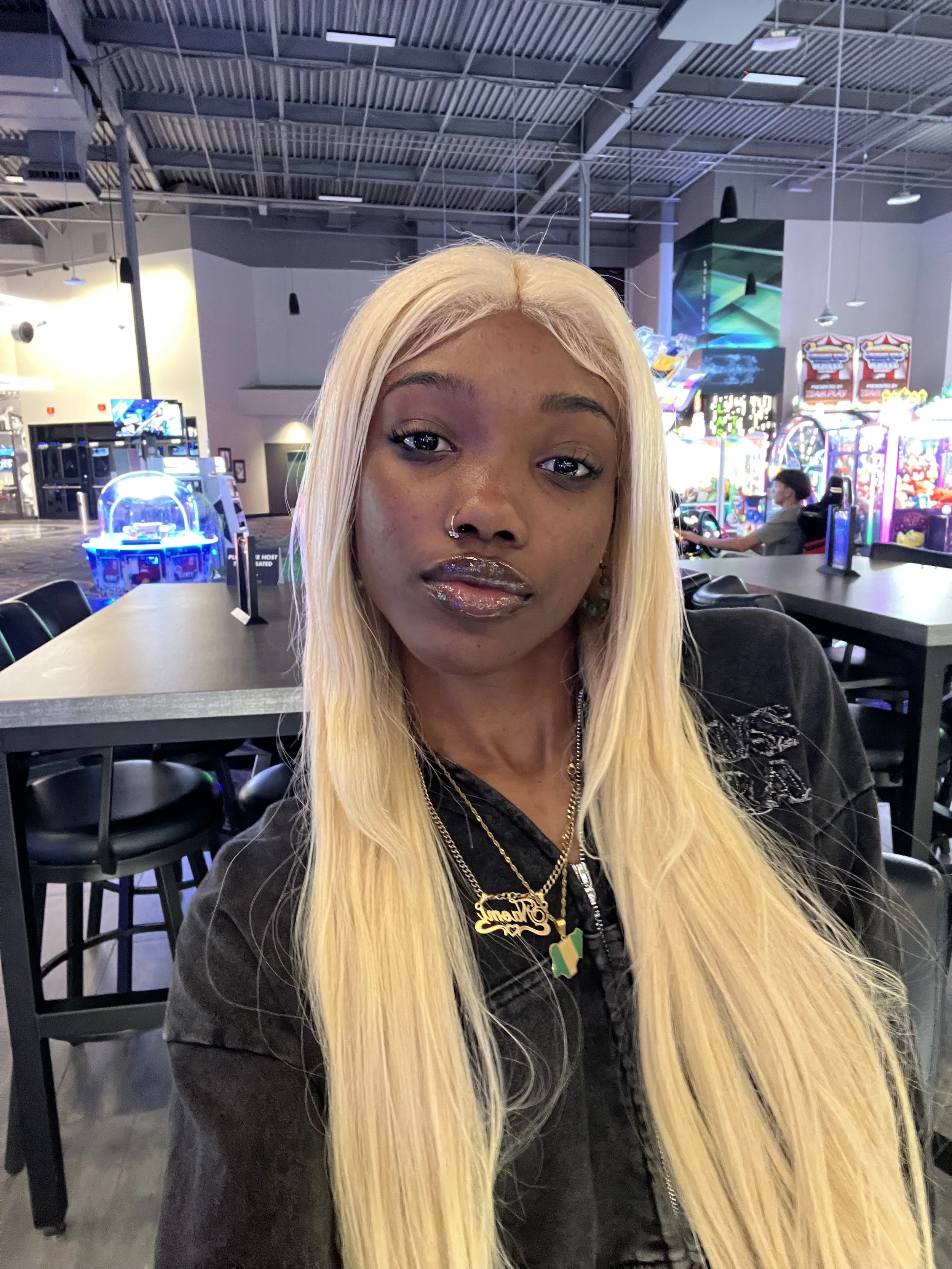 A woman with long blonde hair and glossy lips taking a selfie in an arcade or gaming center with colorful lighting and arcade machines in the background.