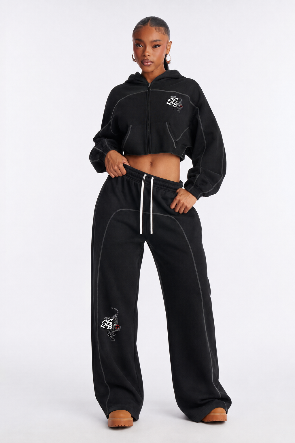 A young woman in a black cropped hoodie and matching sweatpants with white stitching and tiger embroidery, standing against a plain white background.