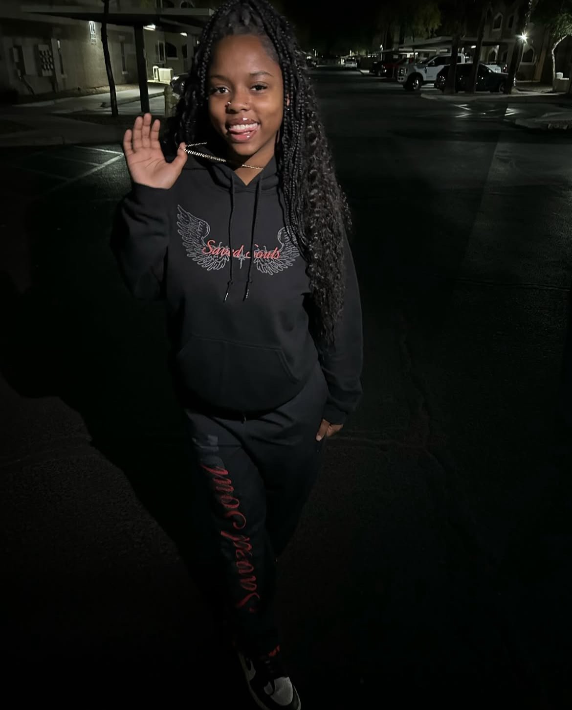 A young woman with long braided hair, wearing a black hoodie and black pants with red writing, standing outdoors at night in a parking lot and smiling at the camera.