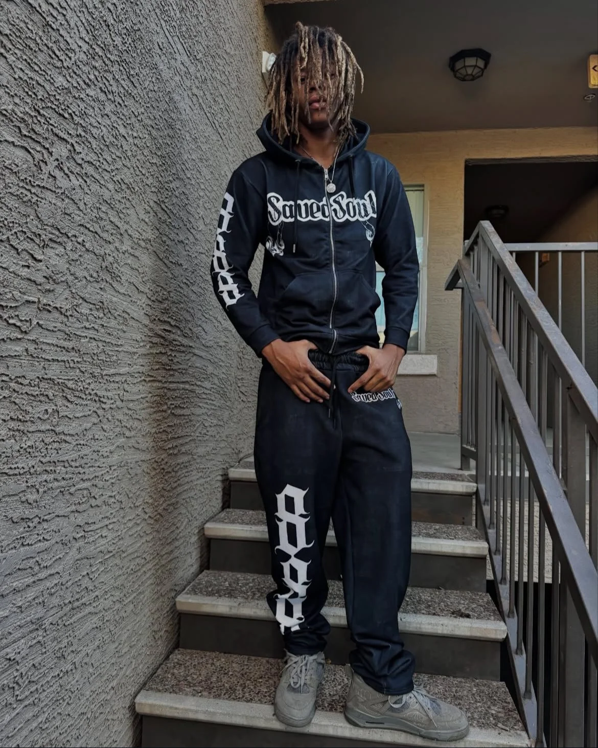 A young man with blonde dreadlocks stands on outdoor stairs, wearing a black zip-up hoodie with white text and black sweatpants with white text, and gray sneakers.