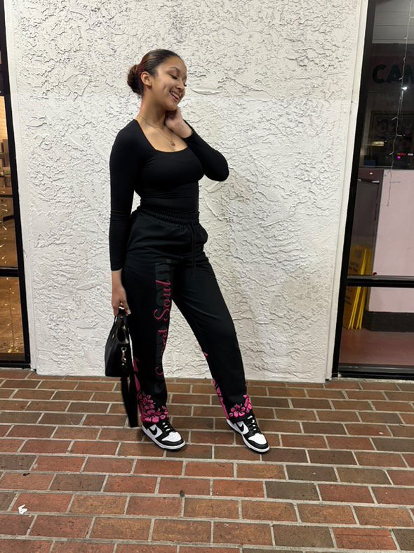 Young woman standing outside against a textured white wall, smiling, wearing a black long-sleeve shirt, black sweatpants with pink floral designs and text, black and white sneakers, holding a black handbag.