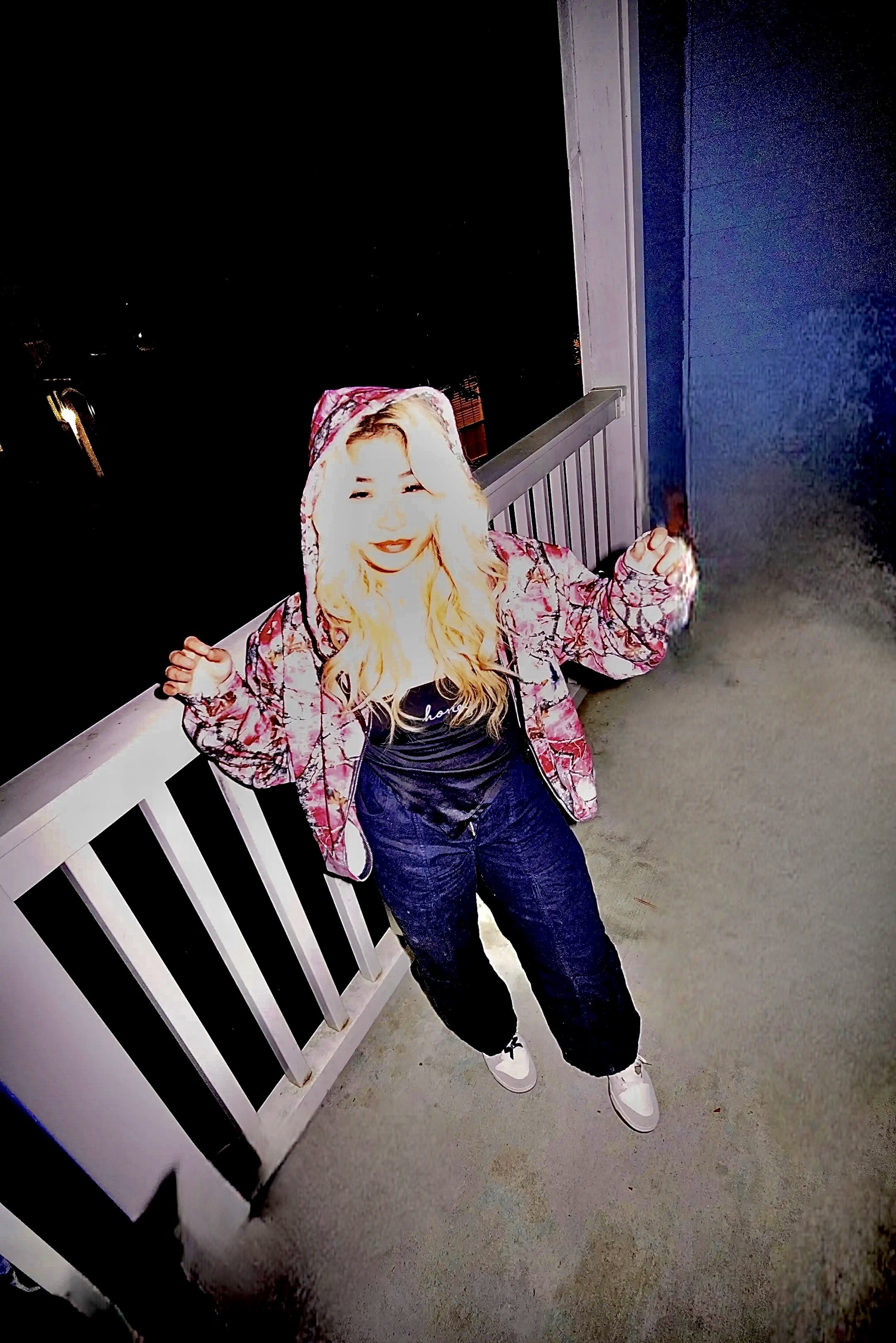 A person standing on a porch at night, wearing a hooded jacket with a pink and white pattern, black clothing underneath, and white shoes. The person's face appears overexposed and light, with long blonde hair, and is smiling with arms raised.