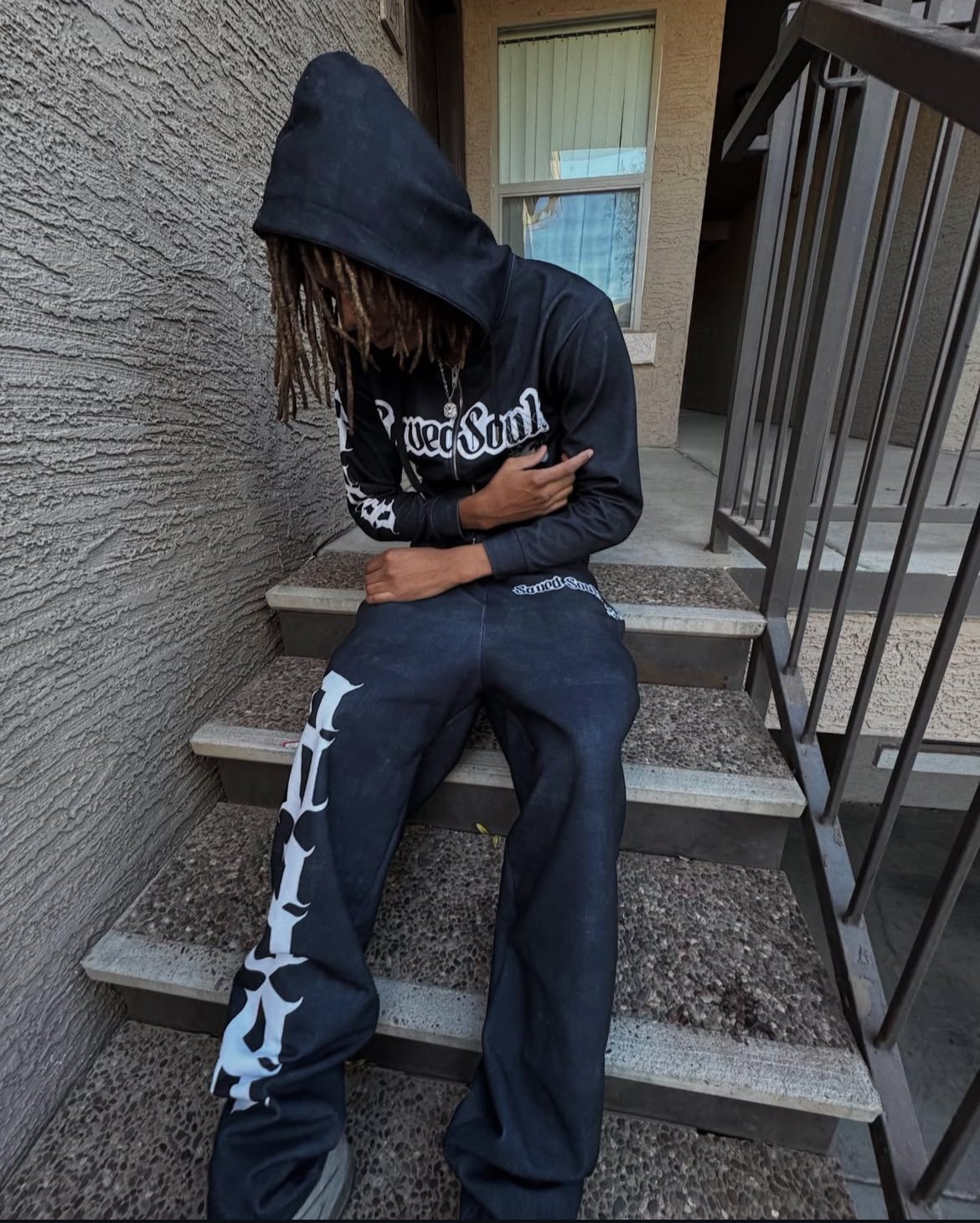 A person wearing a black hoodie with the hood up, black pants with white lettering, and a chain necklace, sitting on outdoor stairs with arms crossed and head down, facing a wall.