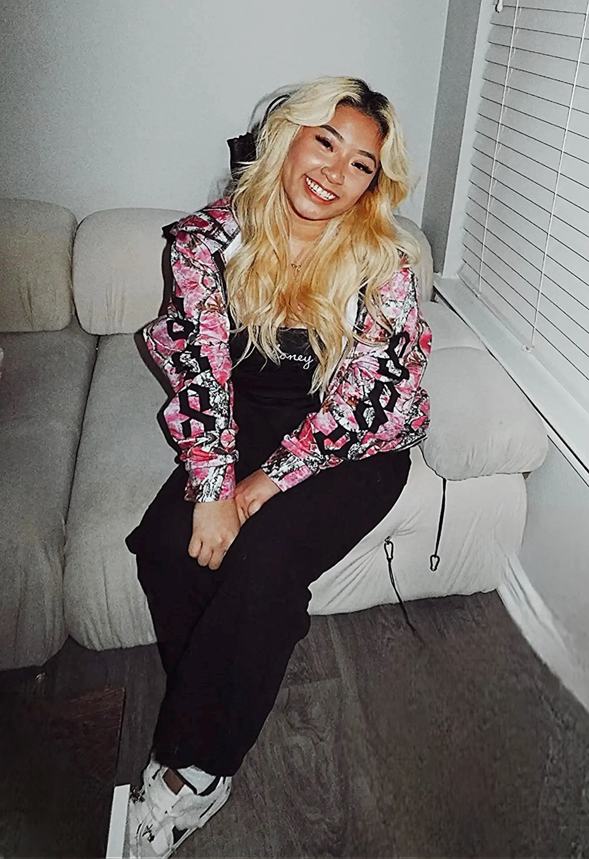 A blonde woman sitting on a beige couch in a living room, smiling at the camera. She is wearing a black shirt, black pants, a colorful patterned jacket, and white sneakers.