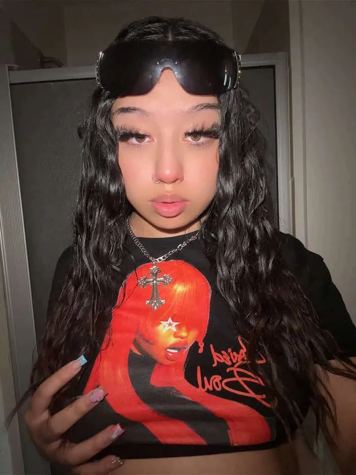 A young woman with long, wavy dark hair, sunglasses on her head, and a nose piercing, wearing a black graphic t-shirt featuring a woman with a star over her eye and a cross necklace.