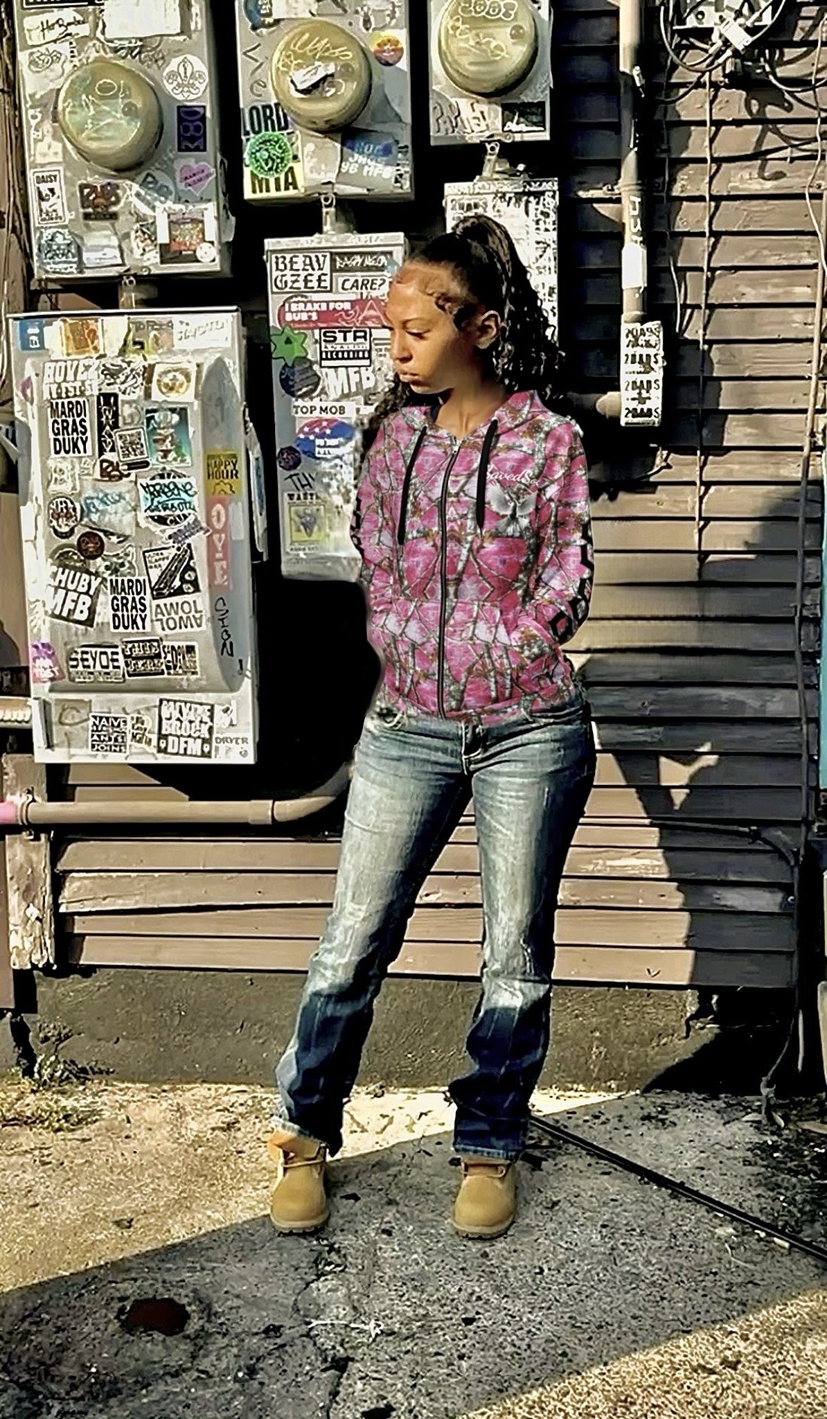 A young woman with braided hair wearing a pink camo hoodie, distressed jeans, and tan boots standing outdoors in front of a wall covered with stickers and a gray rolling shutter.