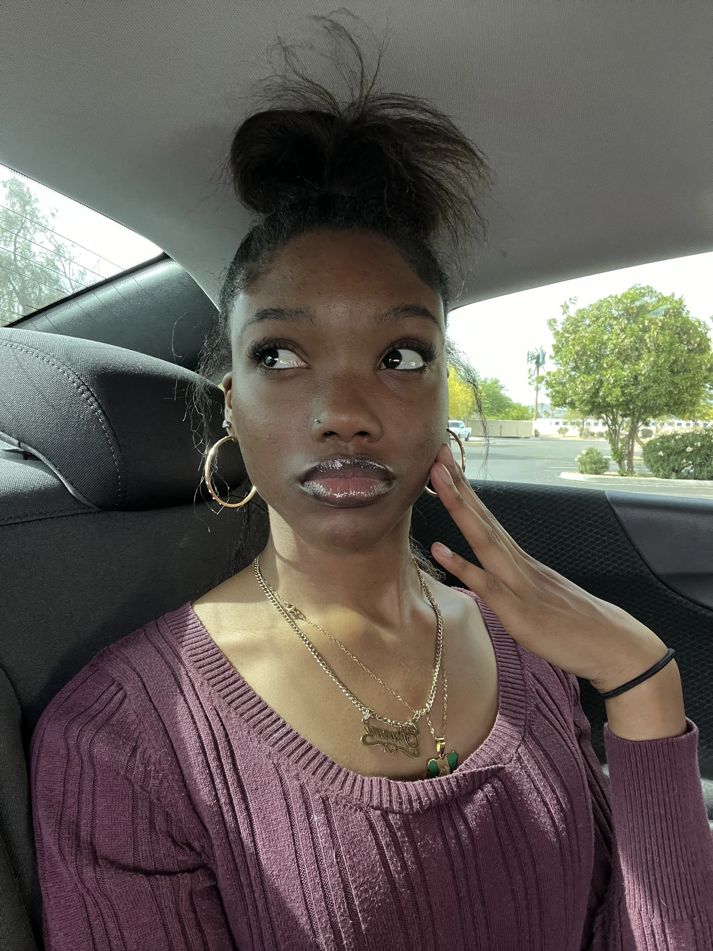 A young woman sitting in a car's back seat with her hair styled in a high, messy bun, wearing a purple ribbed sweater, large hoop earrings, layered gold necklaces, and a nose piercing. She is touching her face with her left hand and looking slightly