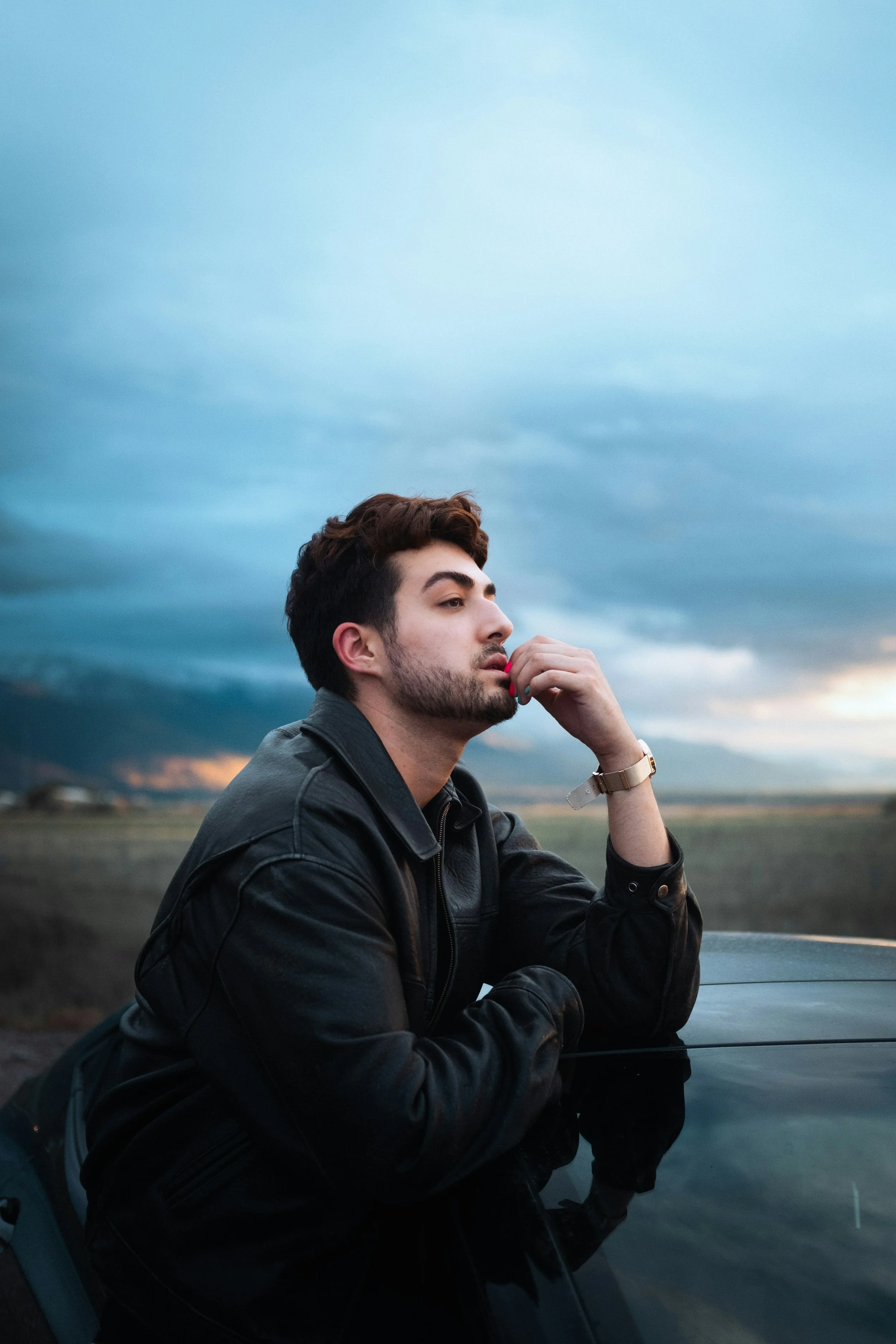 A person with brown hair and a beard, wearing a black leather jacket, sitting outdoors on a car hood, looking thoughtful against a cloudy sky.