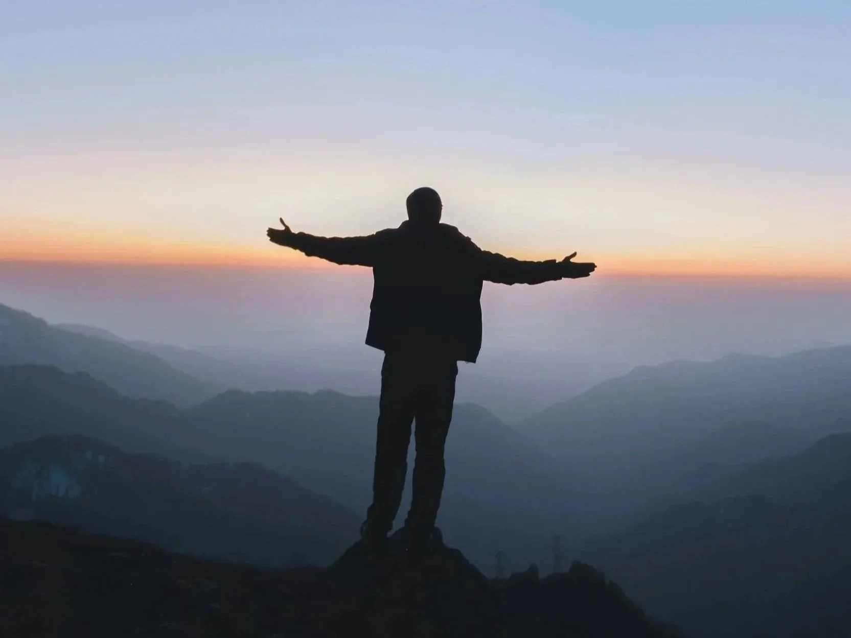 Silhouette of a person standing on a hill with arms outstretched towards a colorful sunset over mountains.