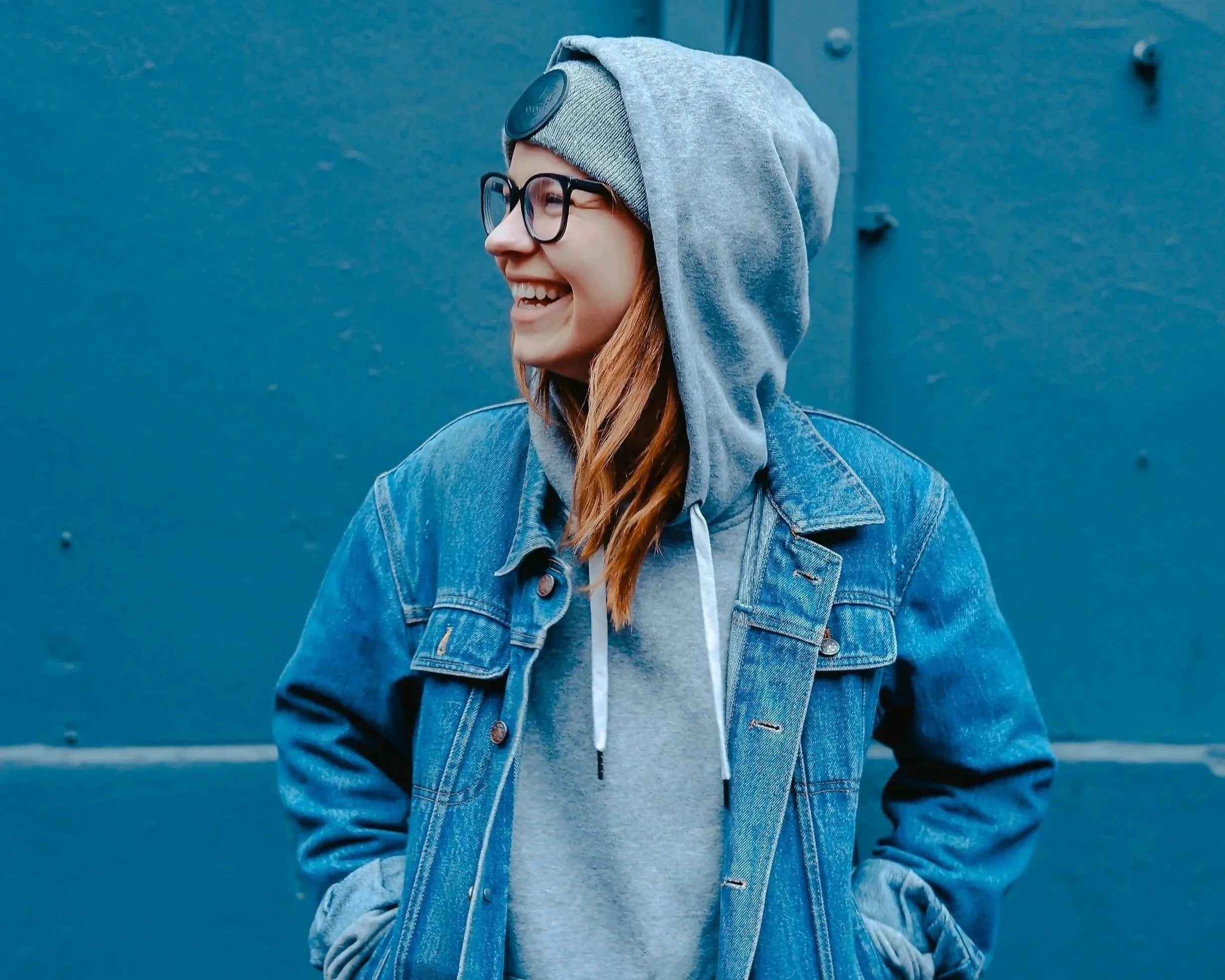 A smiling young person wearing glasses, a gray hoodie, a denim jacket, and a gray beanie, standing against a blue wall.