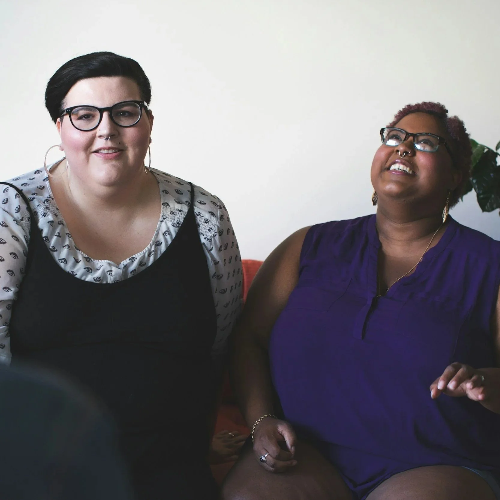 Two neurodivergent people sitting on a couch, smiling and engaging in conversation with a person whose back is facing the camera, indoors with a plant in the background.