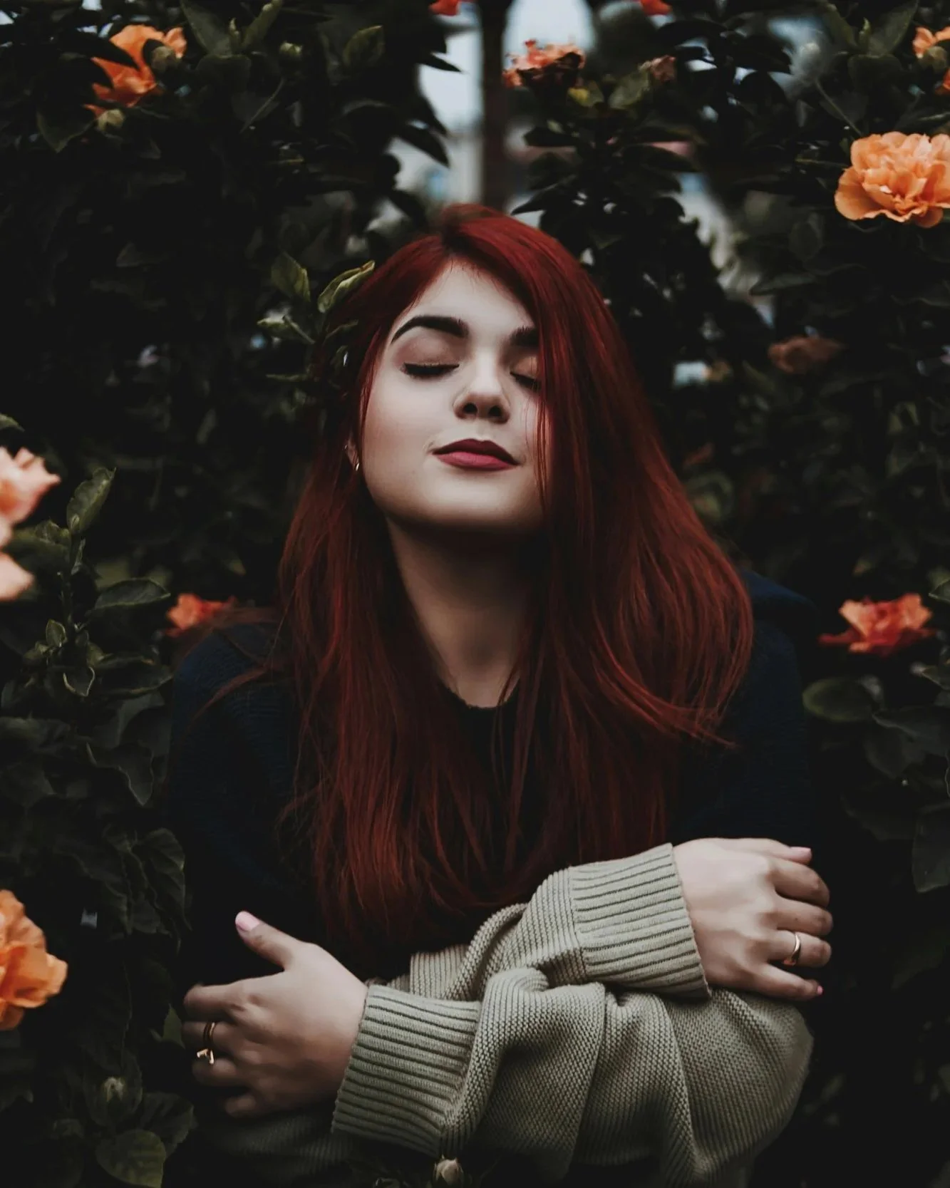 A person with red hair and closed eyes standing among dark green bushes and orange flowers.