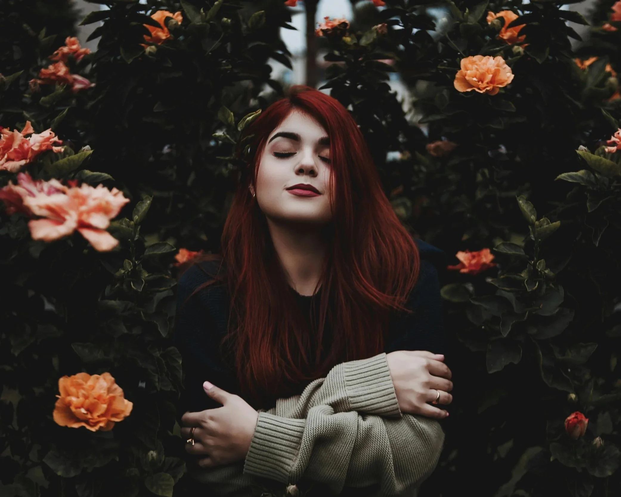 A young person with long red hair and closed eyes standing amidst dark green bushes with peach-colored flowers, their arms crossed and a calm expression.