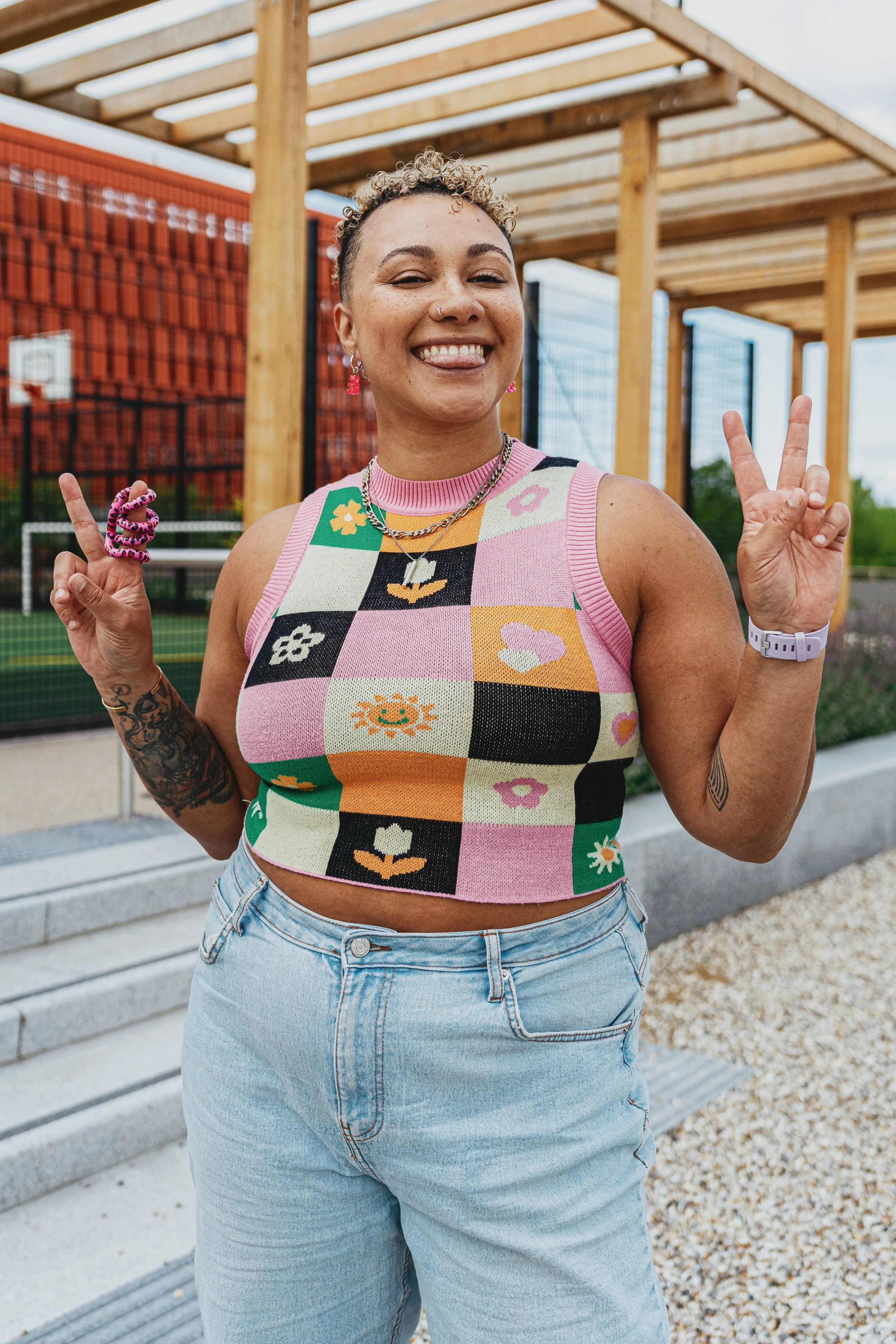 A young person standing outdoors in front of a sports court, smiling and making peace signs with both hands. They are wearing a colorful, patterned sleeveless crop top and light-wash jeans.