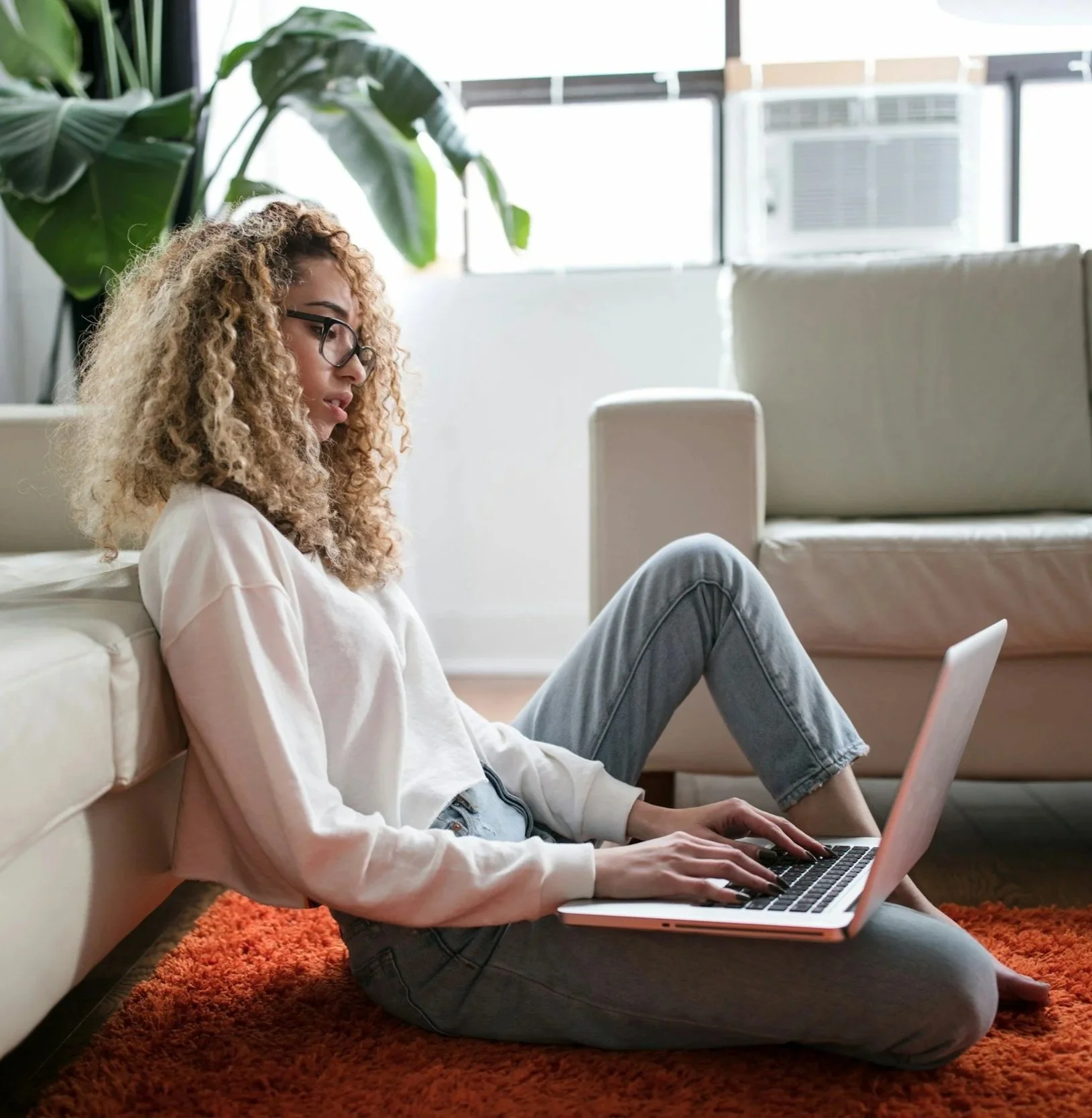 A young person with curly hair and glasses sitting on the floor with a laptop, leaning against a couch in a bright living room with large windows and a potted plant.