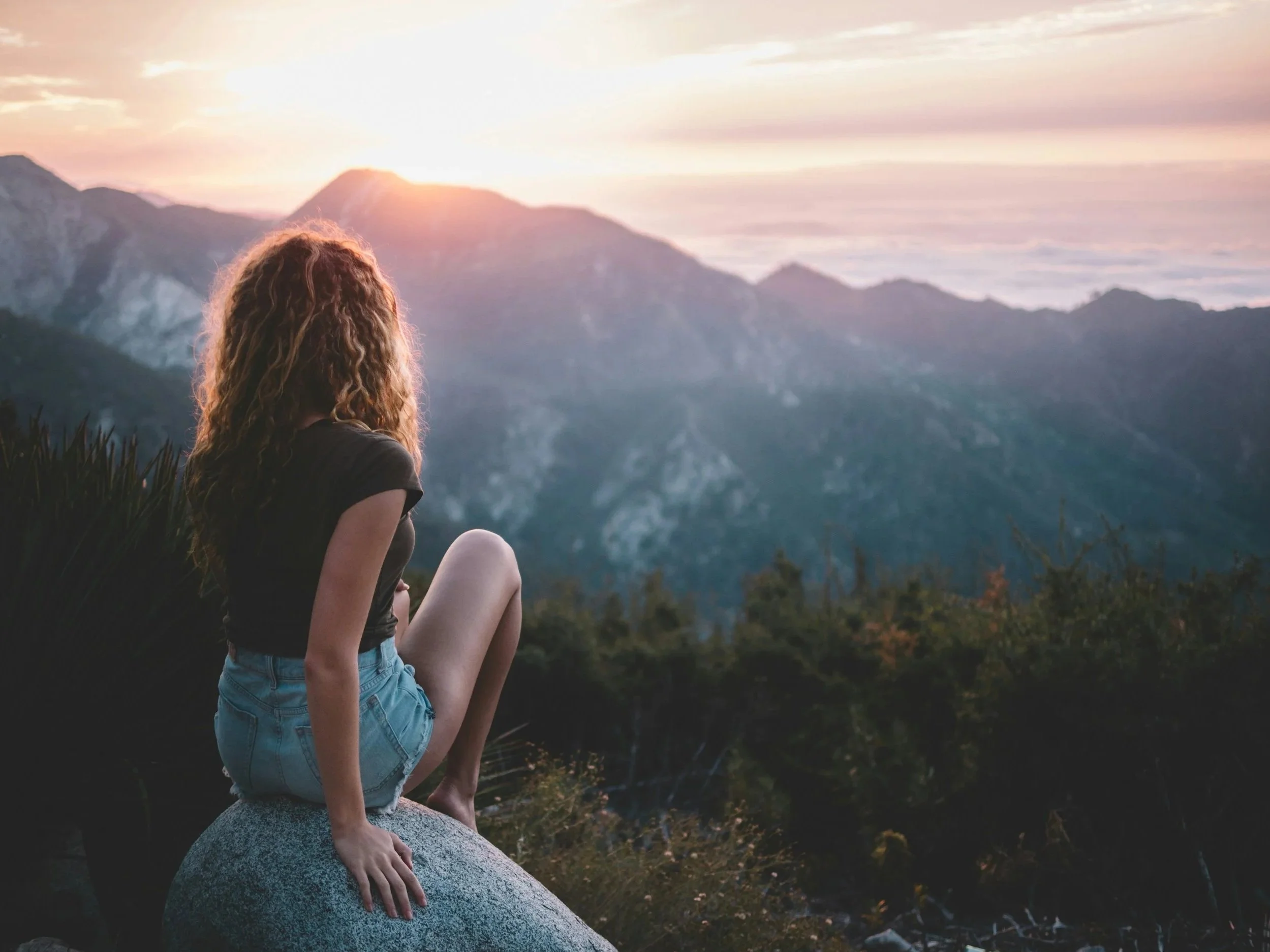 A young person with curly hair sitting on a rock, gazing at a sunset over mountain peaks and the ocean.