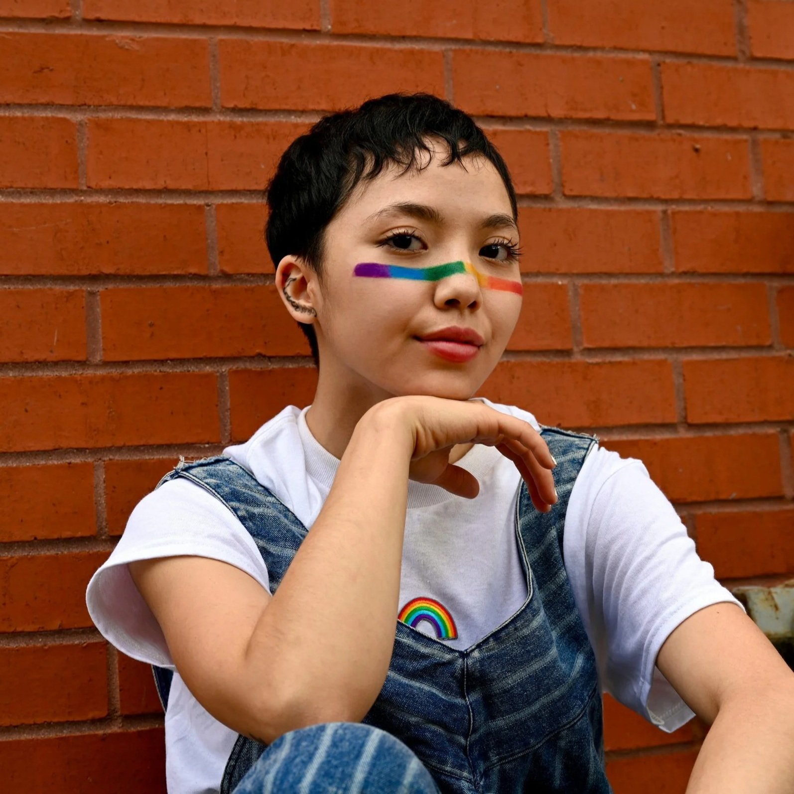 A person with short dark hair and light skin, sitting against a red brick wall, with rainbow face paint across their cheekbones and nose, and a rainbow sticker on their denim overalls.