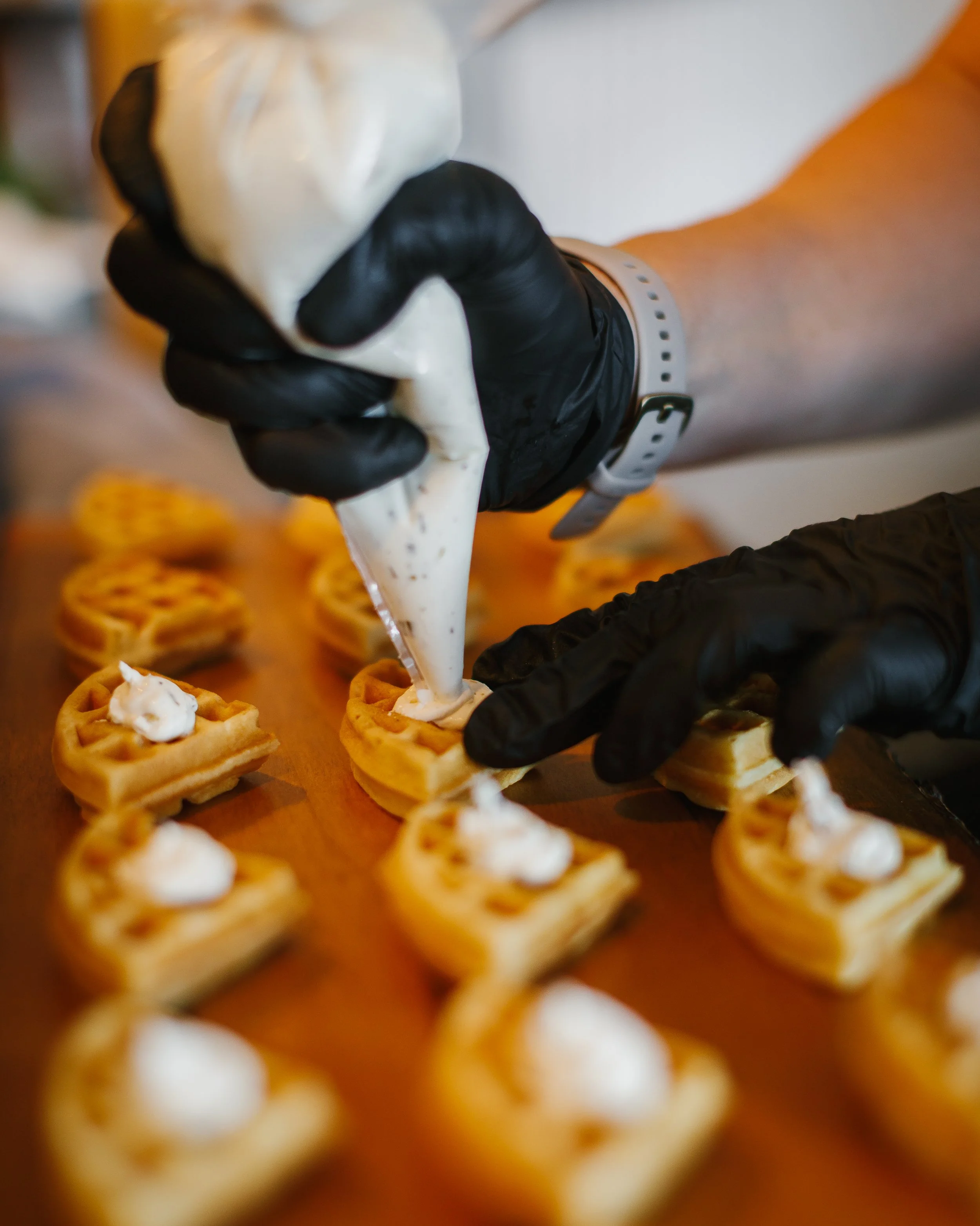 Person wearing black gloves piping cream onto small waffles arranged on a wooden surface.