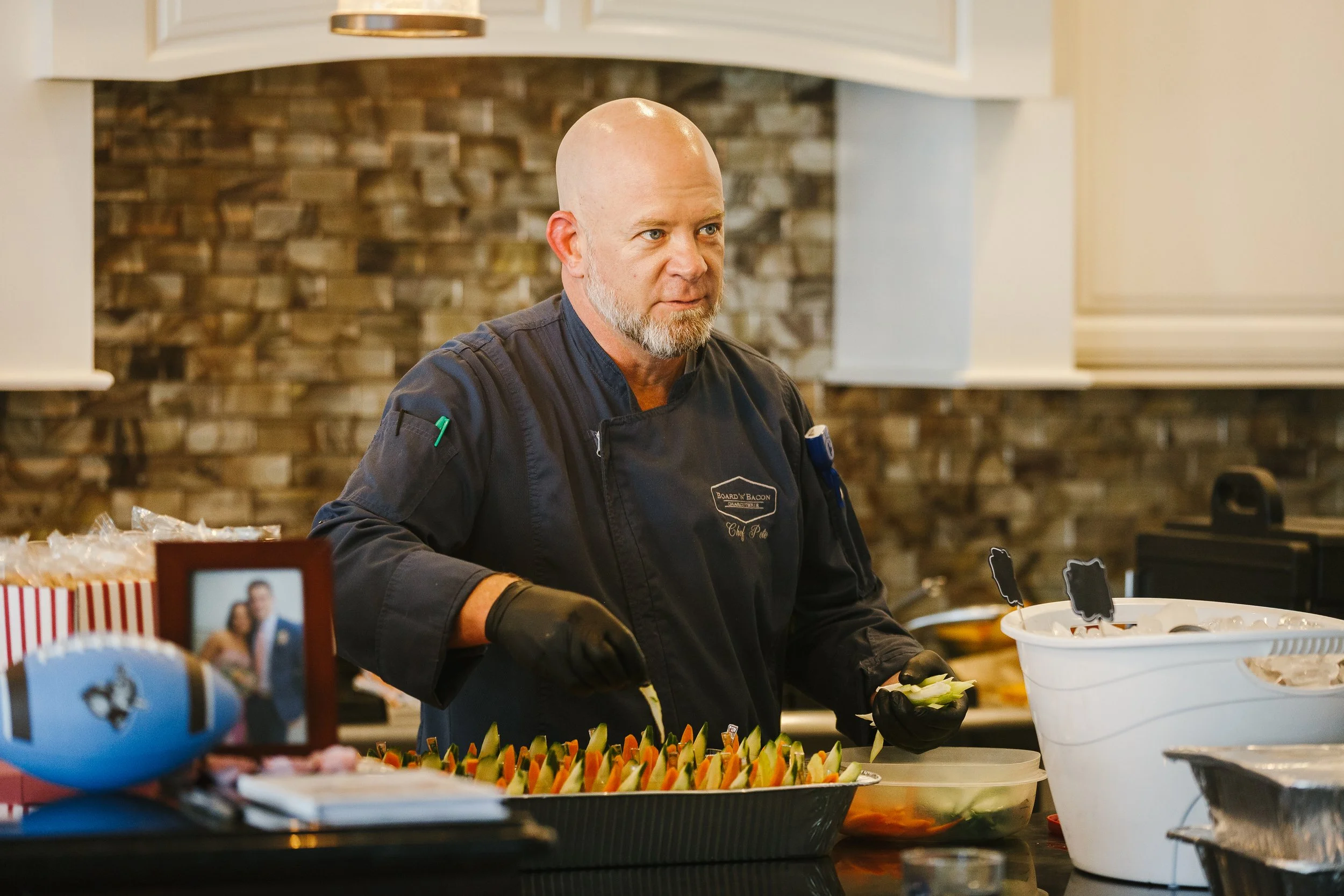 A chef with a bald head and gray beard wearing a dark chef's coat and black gloves preparing food in a kitchen.