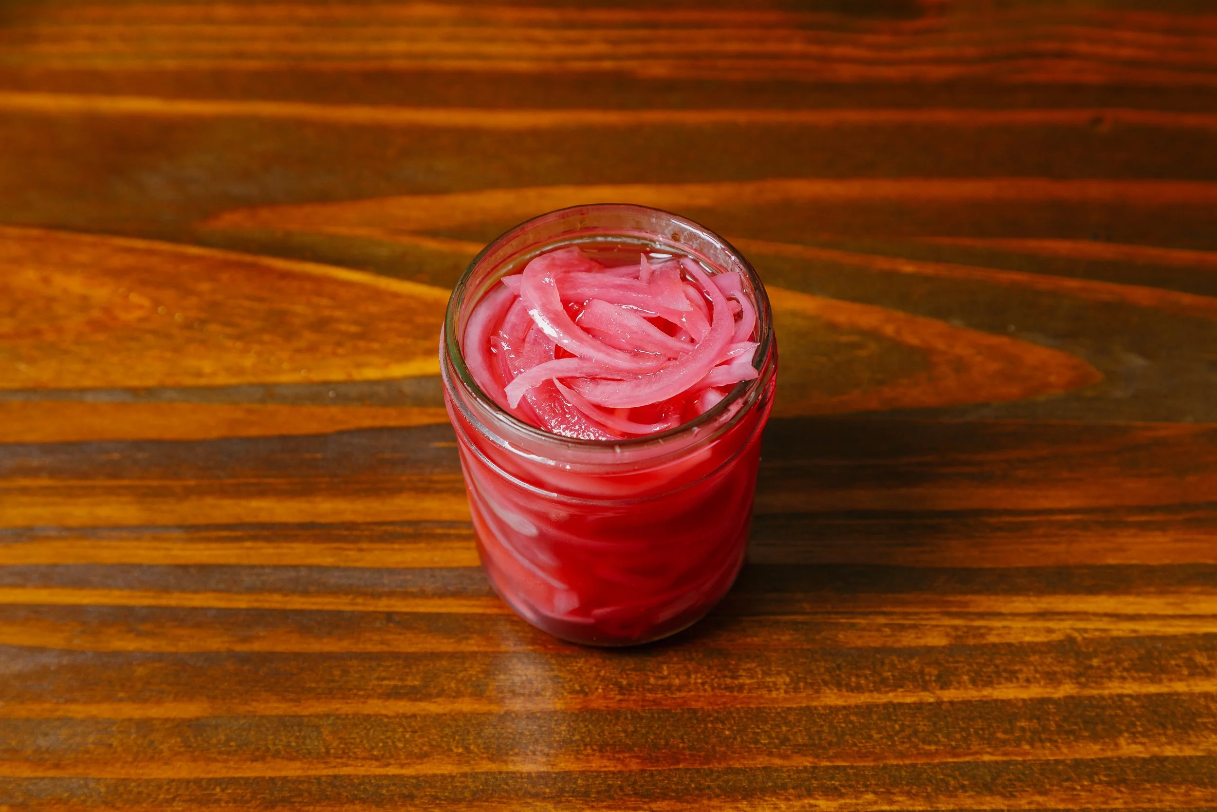 A glass jar filled with pink pickled onion slices on a wooden surface.