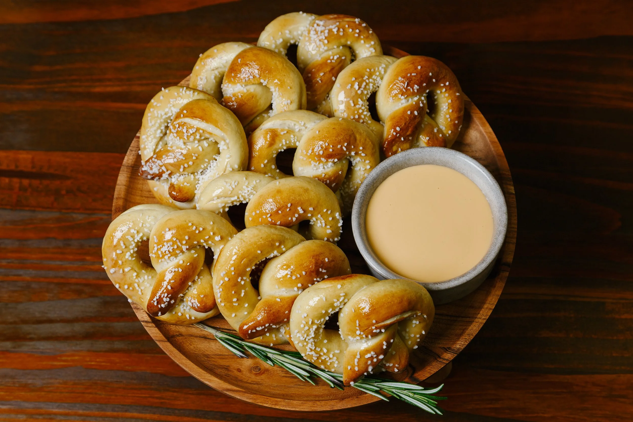 A round wooden platter filled with small pretzels sprinkled with coarse salt, accompanied by a bowl of cheese sauce, garnished with a sprig of rosemary on a dark wooden table.