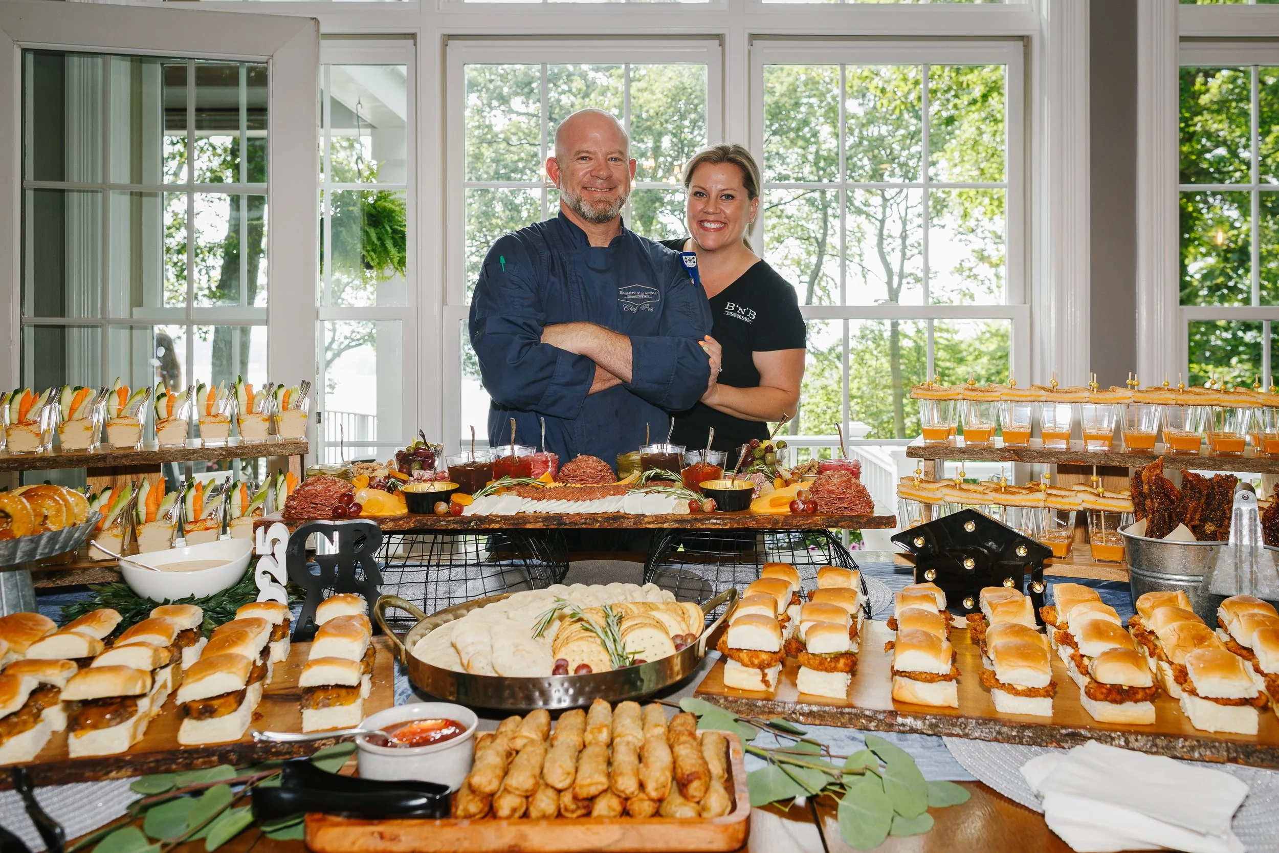 Two people, a man in a chef's jacket and a woman in a black shirt, stand behind a table filled with various appetizers, sandwiches, and desserts, with a large window and green trees in the background.