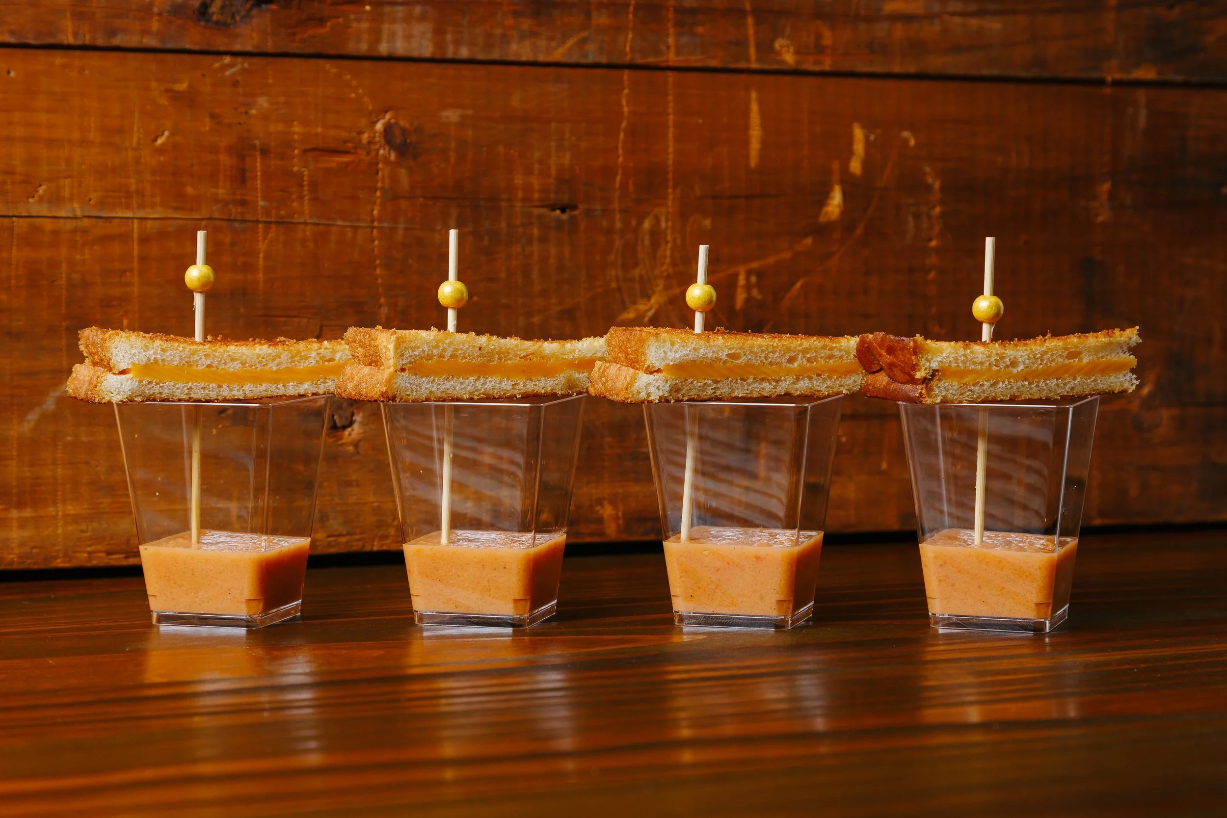 Four glasses of a creamy orange beverage with toasted sandwich slices on top, each with a decorative toothpick, arranged on a wooden surface against a wooden background.
