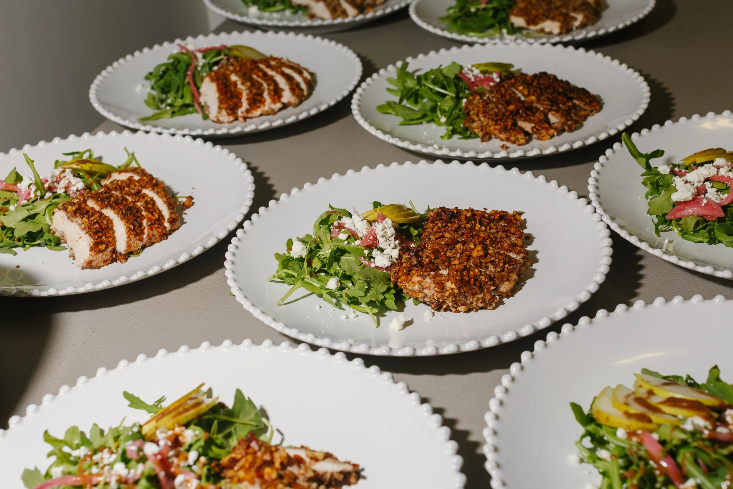 Multiple white plates with breaded, fried chicken or meat cutlets and mixed green salads with cherry tomatoes, cheese crumbles, and dressing, arranged on a gray surface.