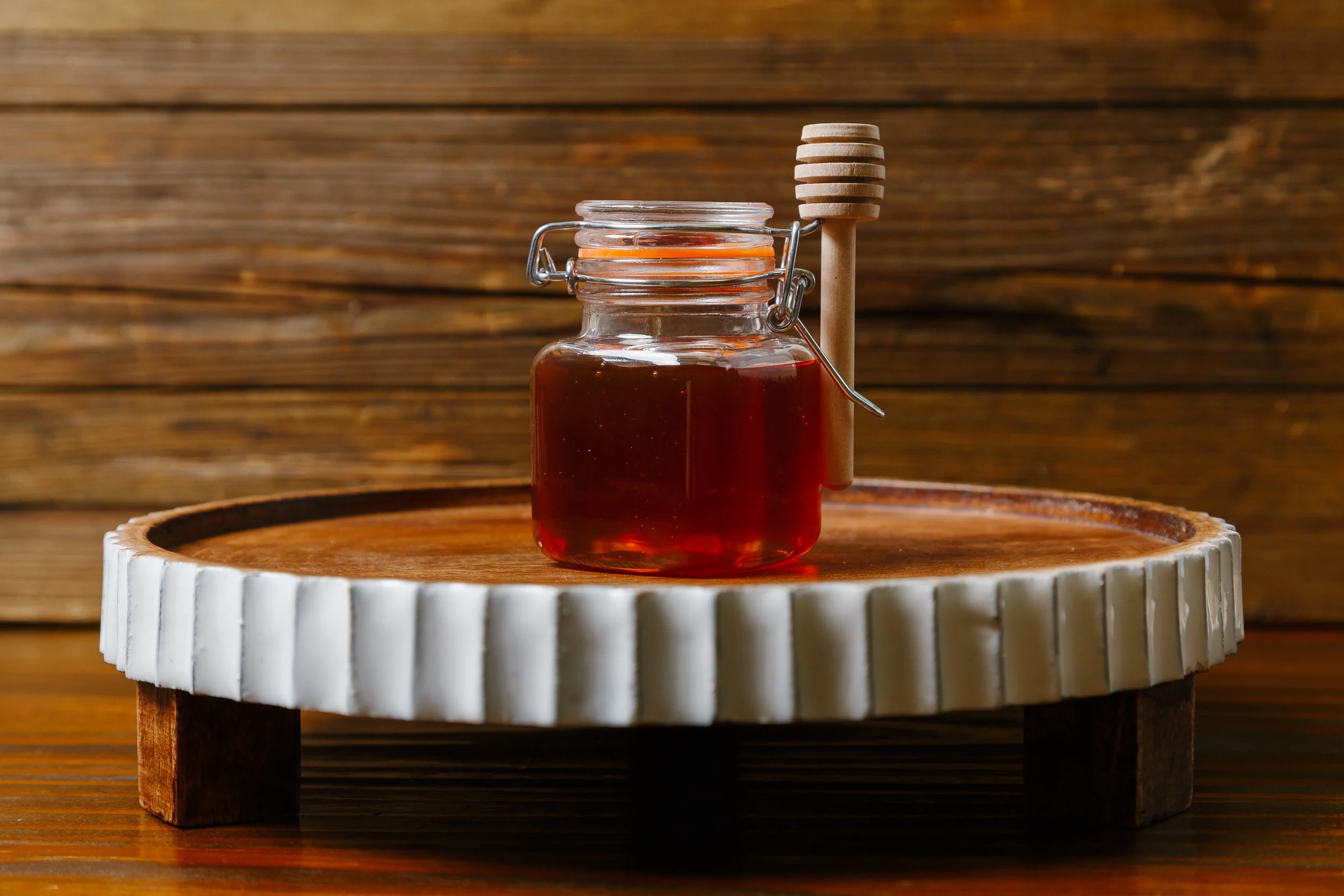 A small glass jar filled with dark red honey on a white-rimmed wooden tray, with a honey dipper hanging beside it, against a wooden background.