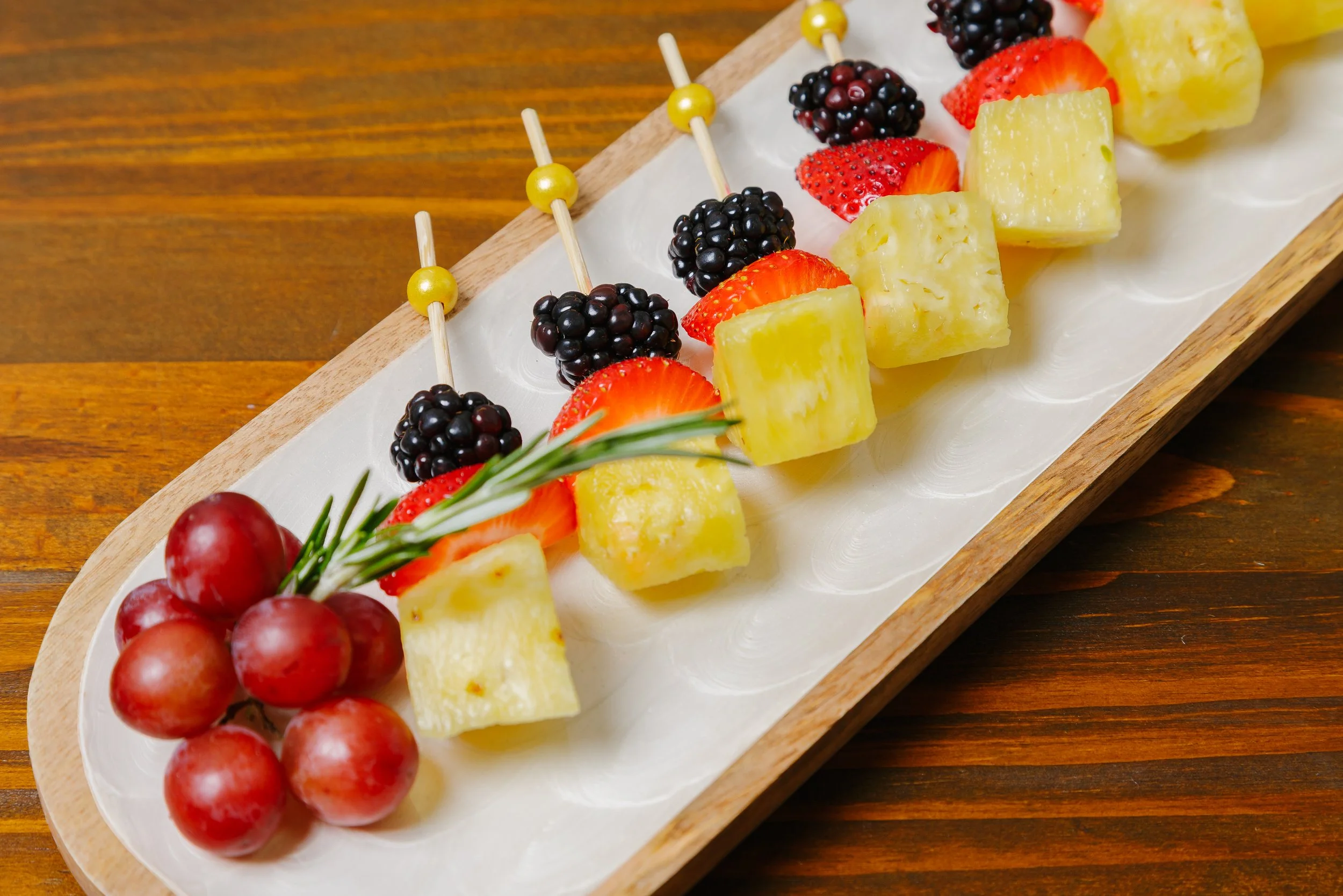 A wooden serving board with skewers of cheese, strawberries, blackberries, and grapes, garnished with a sprig of rosemary.