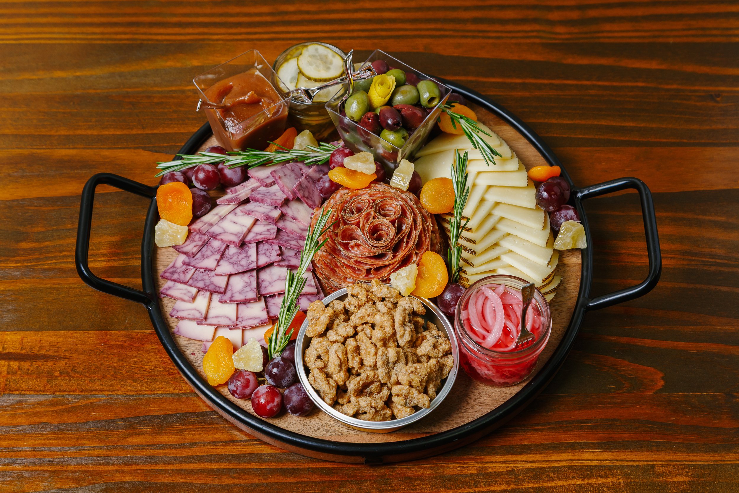 Charcuterie board with sliced meats, cheeses, olives, pickles, dried fruits, and garnished with sprigs of rosemary on a wooden table.