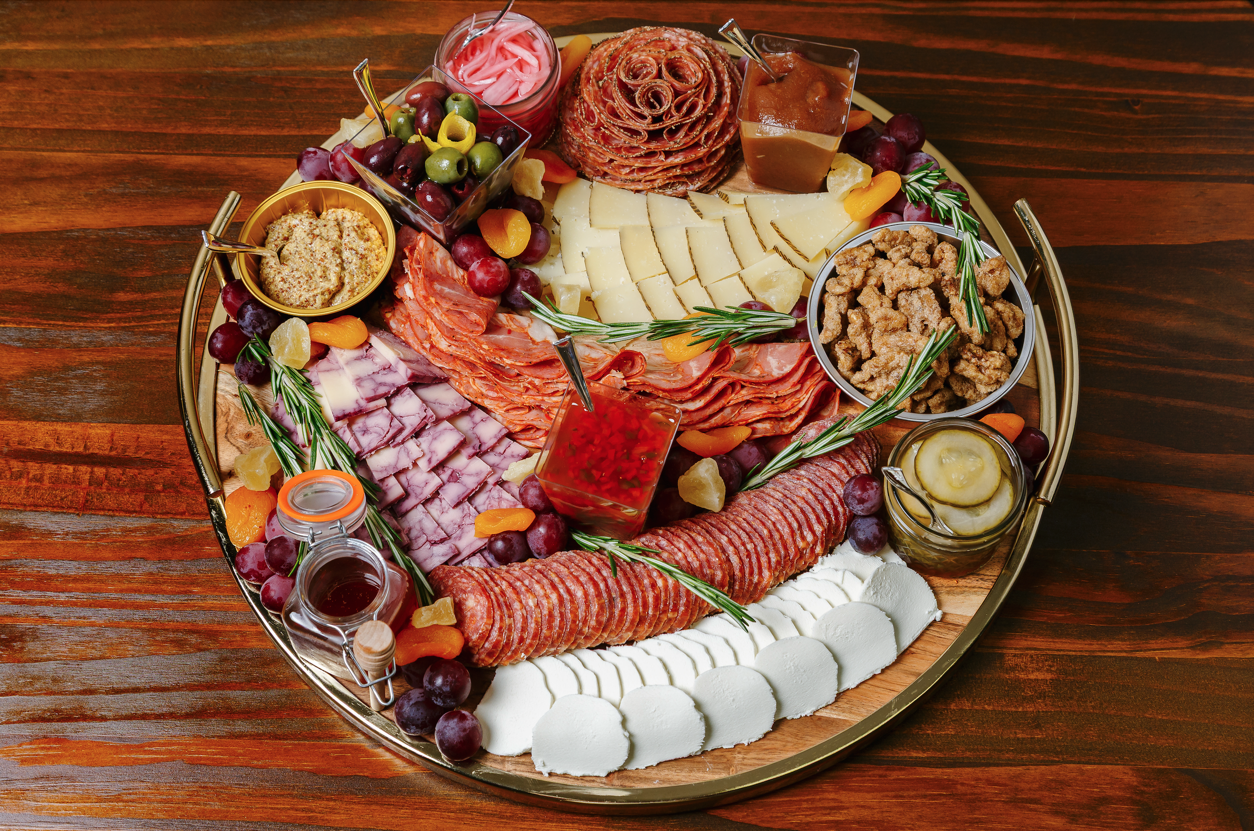 A round meat and cheese platter with various sliced meats, cheeses, fruits, pickles, and condiments on a wooden table.
