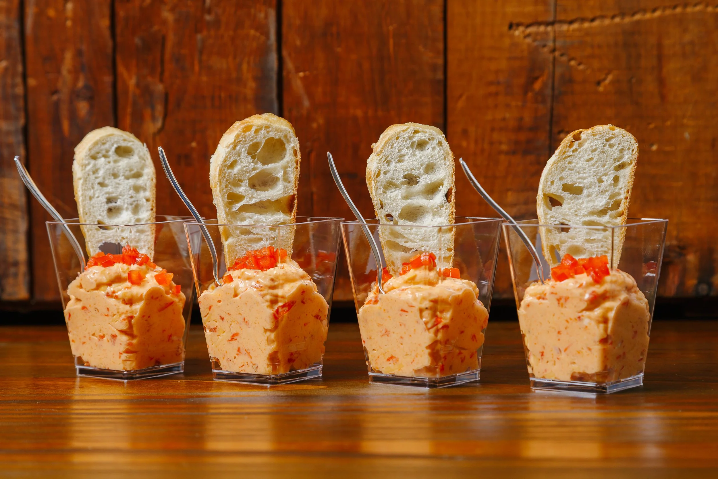 Four small glasses filled with a creamy orange-colored dip, garnished with small red pieces, each with a slice of toasted baguette.