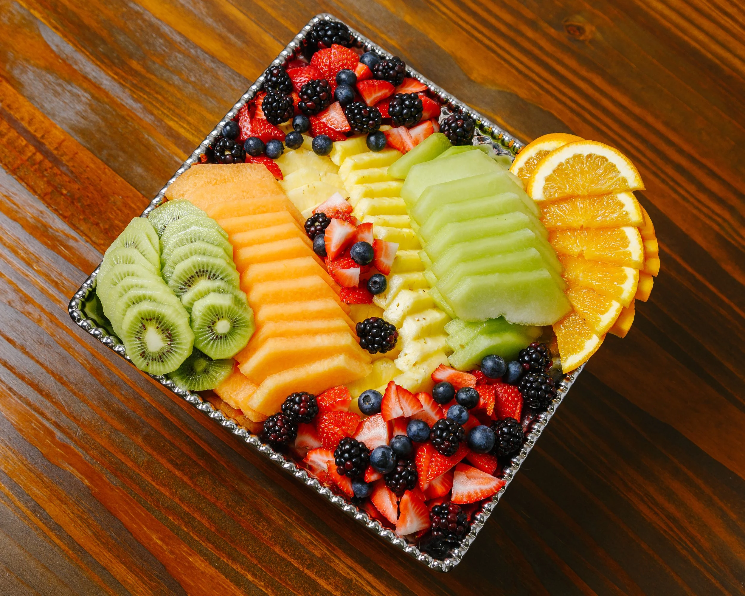 Assorted fresh sliced fruits including kiwi, cantaloupe, honeydew, orange, strawberry, blackberry, and blueberry in a decorative tray on a wooden table.