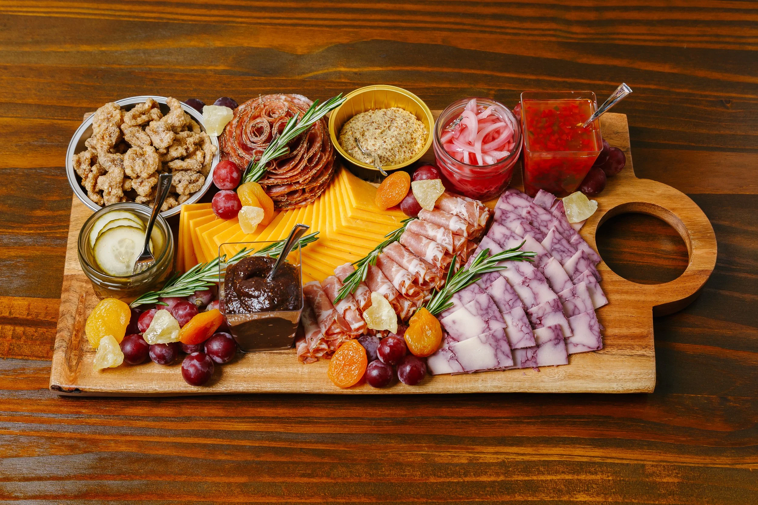 A charcuterie board with cheeses, cured meats, grapes, pickles, dried fruit, nuts, and various dipping sauces on a wooden table.