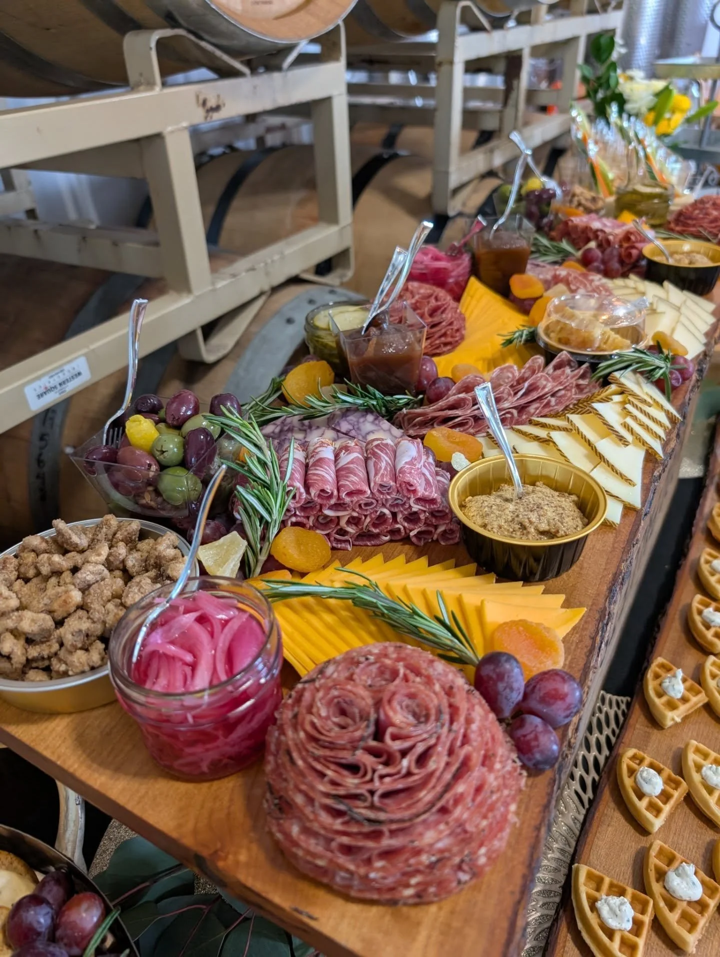 A charcuterie board with sliced meats, cheese, fruit, pickles, and condiments, arranged on a wooden surface with wine barrels in the background.