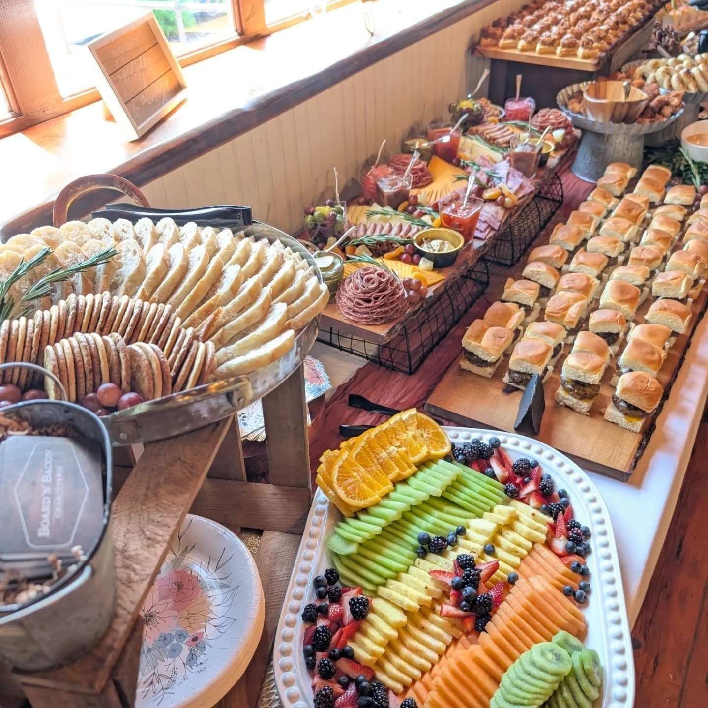 A table with an assortment of food including sliced bread, various pastries and cookies, a colorful fruit platter with sliced melons, strawberries, blackberries, blueberries, and oranges, and a variety of finger sandwiches with different fillings. Th