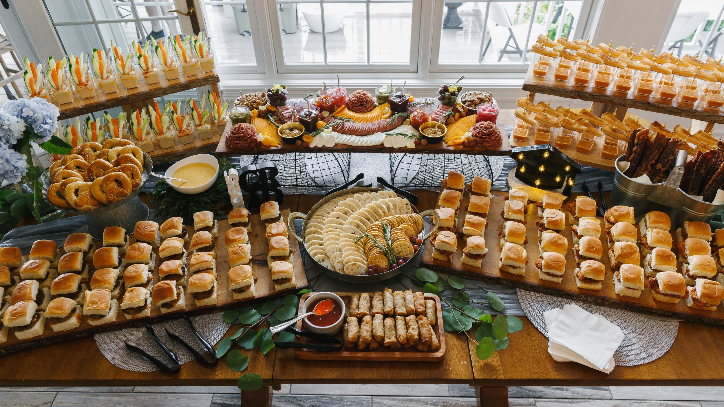 A large buffet table filled with various appetizers and desserts, including finger sandwiches, cookies, fruit parfaits, and small cakes, arranged on wooden and ceramic serving trays with decorative greenery and flowers.