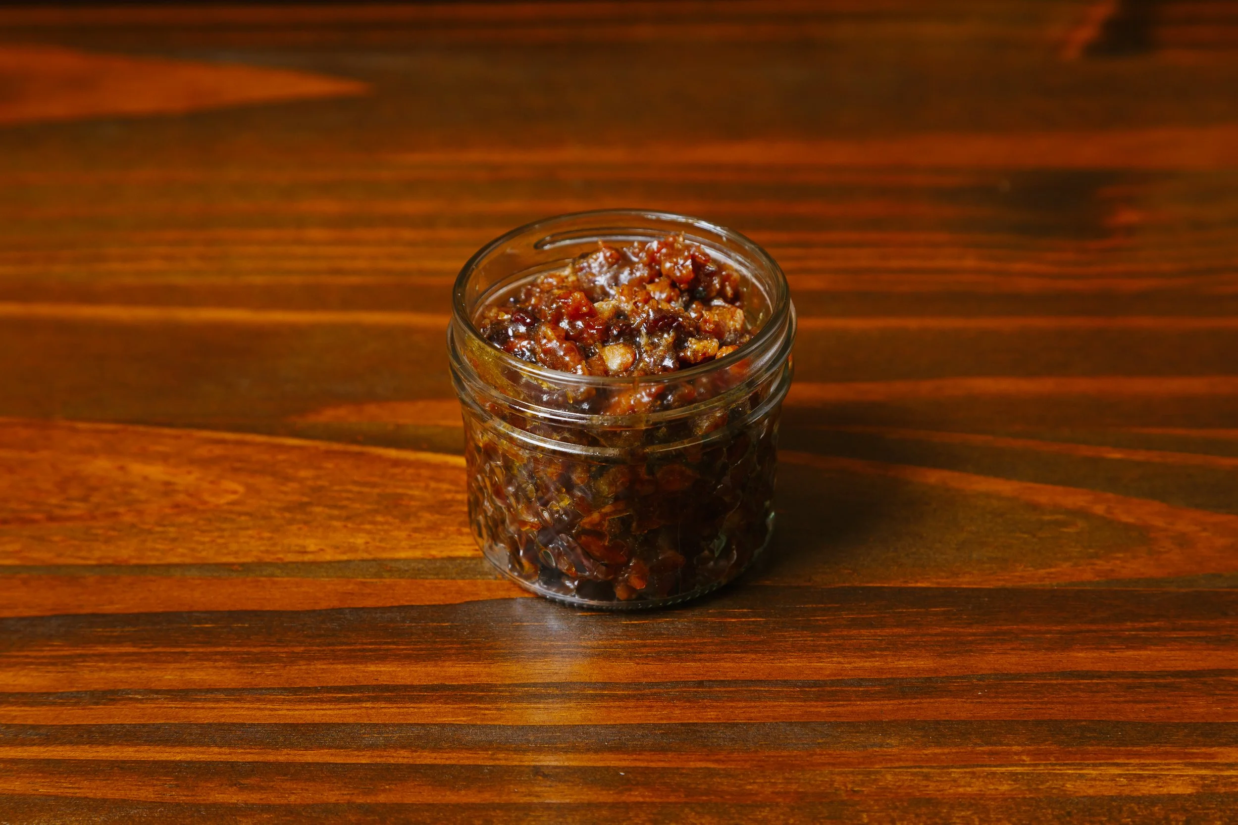 A small glass jar filled with a dark, chunky relish or chutney placed on a wooden surface.