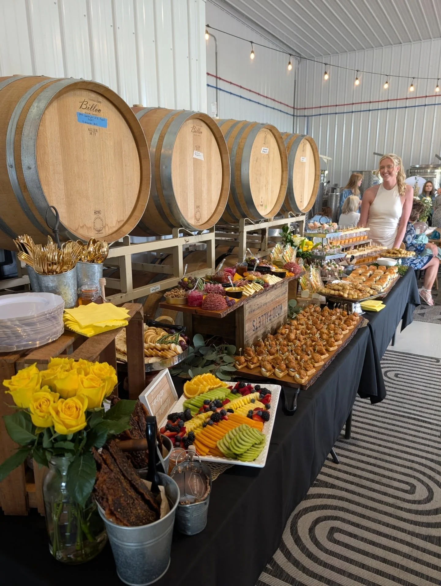 A dessert buffet setup at an event with several wine barrels in the background, and a woman smiling behind the table. The table has plates of fresh fruit, assorted pastries, small desserts, and a decorative bouquet of yellow roses.
