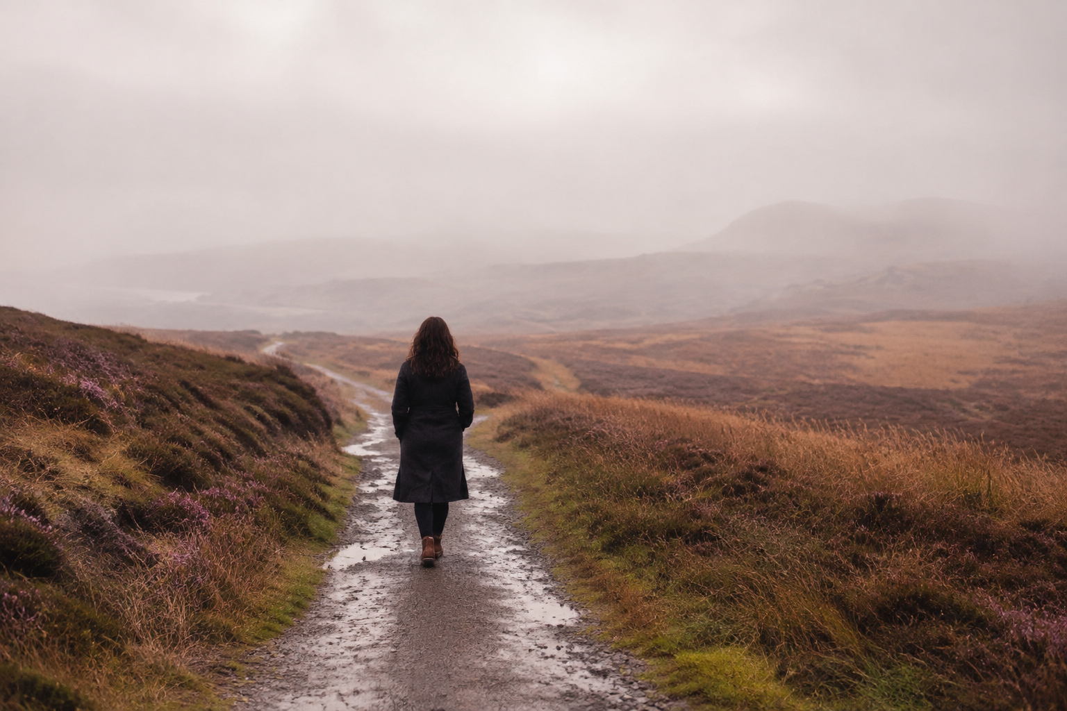 A woman walking alone along a wet countryside path in the mist, reflecting how grief can feel harder as time passes.