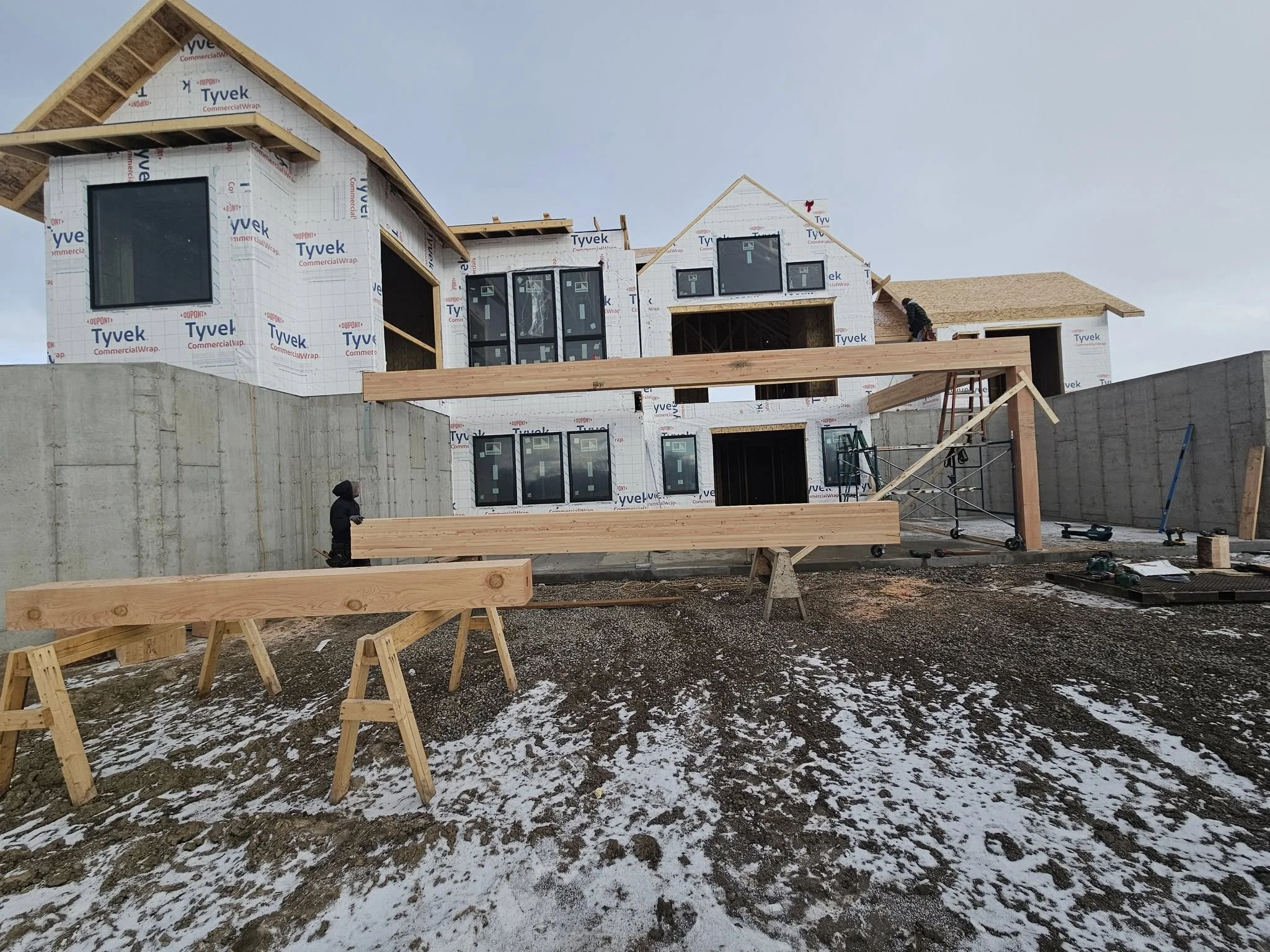 Under construction house with wooden framework, large windows, and a concrete retaining wall with snow on the ground.
