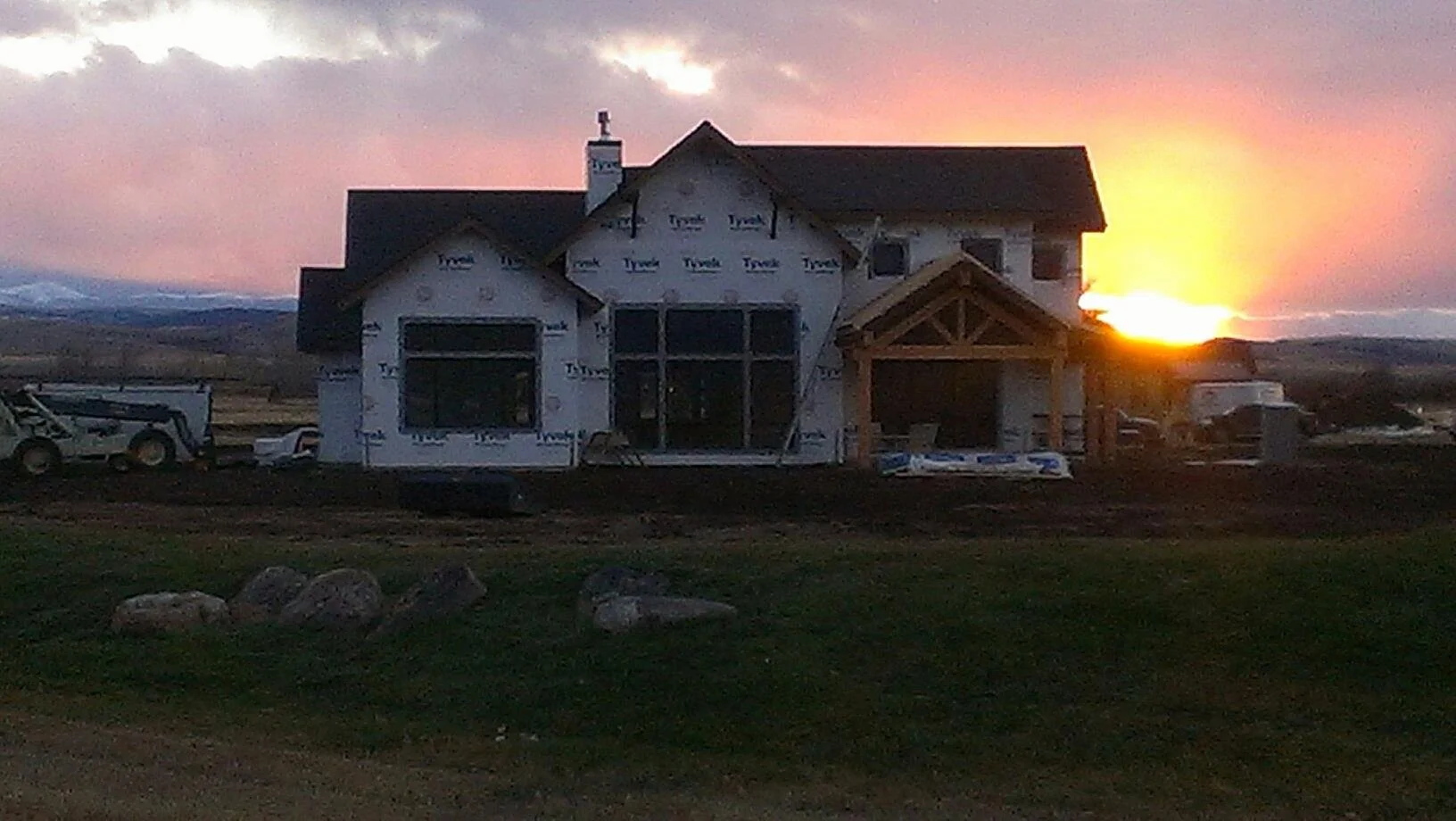 A house under construction at sunset, with construction vehicles and materials nearby and a partially finished exterior.