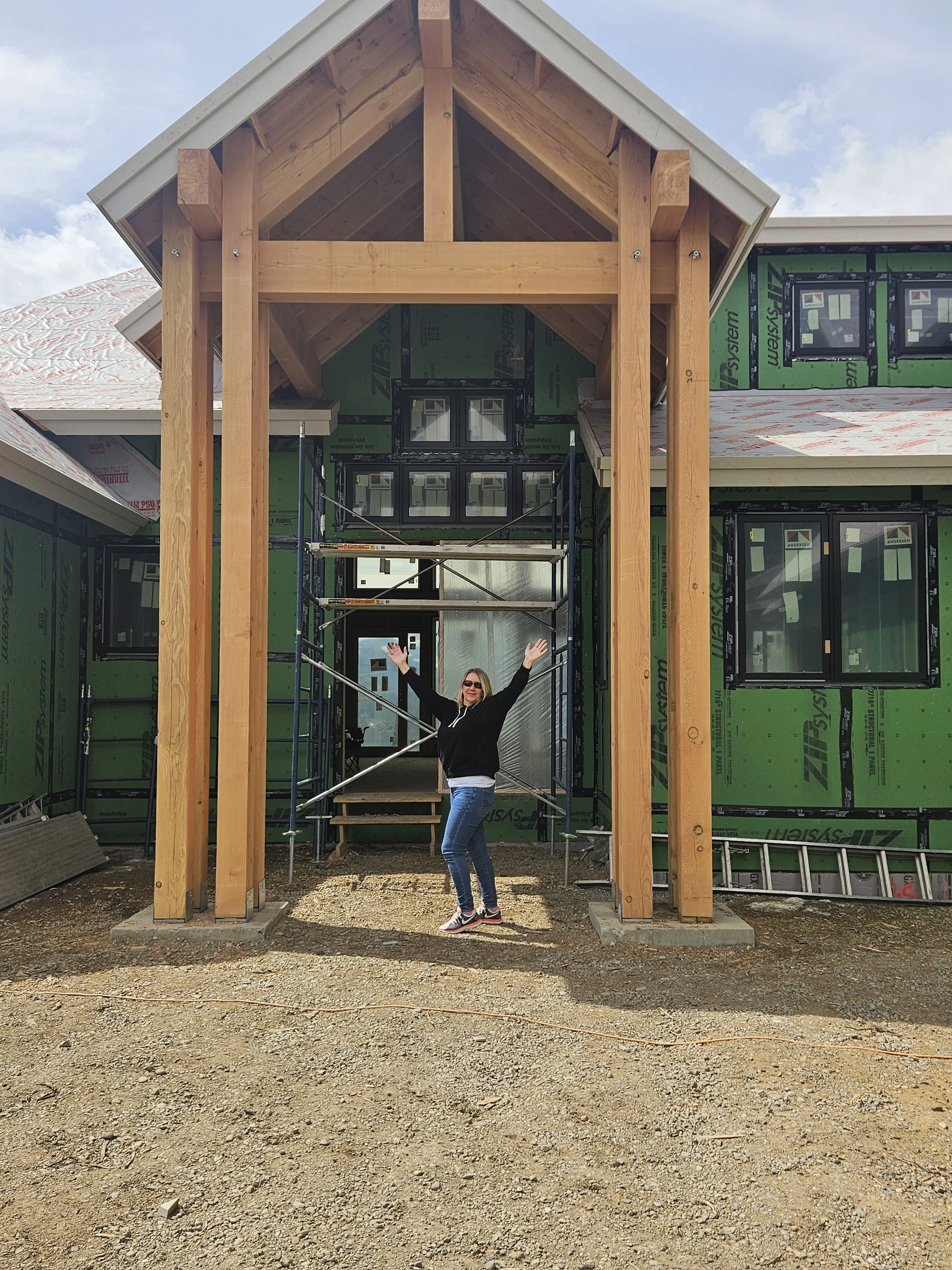 A woman standing in front of a house under construction, with her arms raised, surrounded by scaffolding.