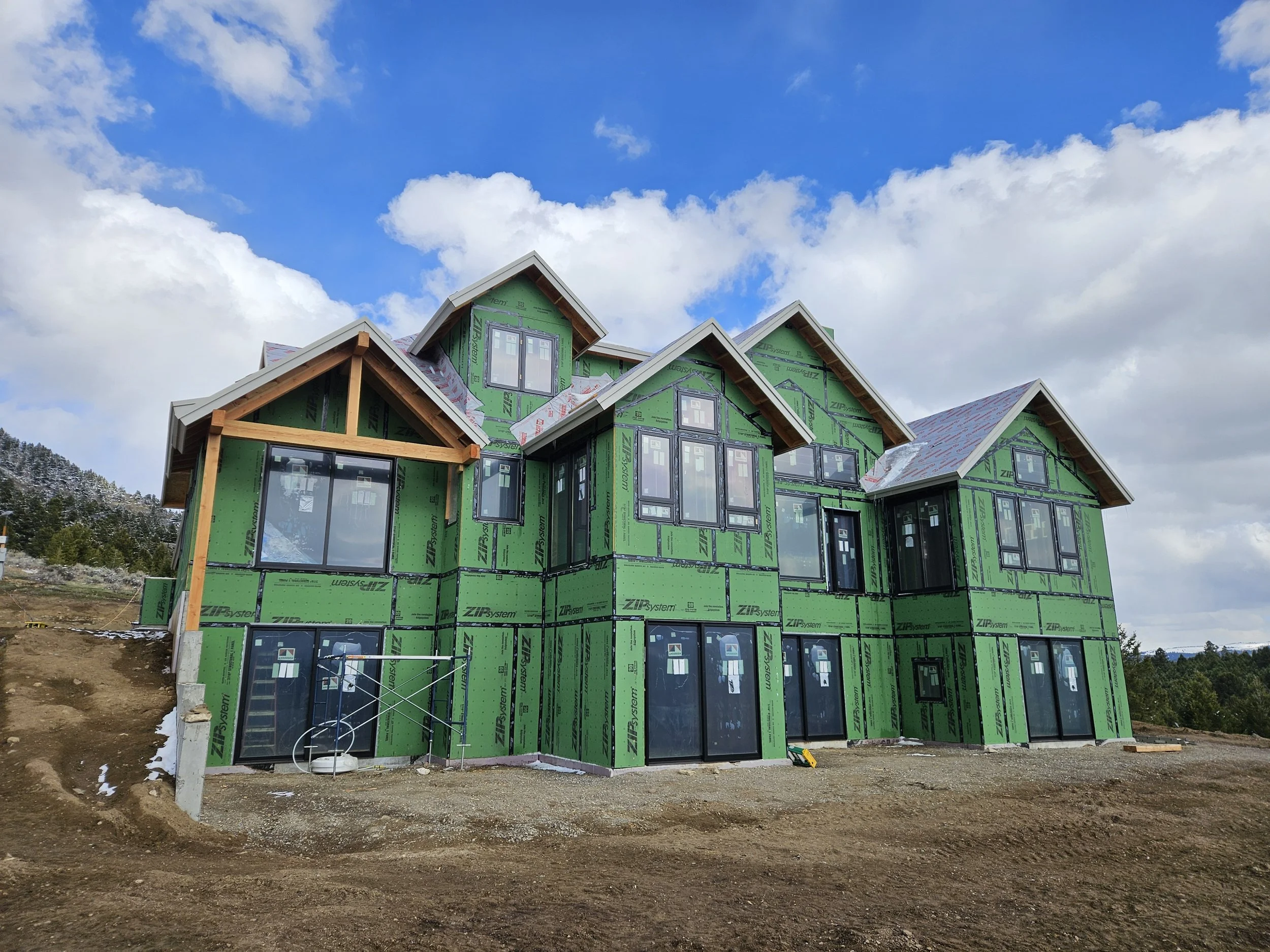 A multi-story house under construction with green exterior sheathing, large windows, and a steep gabled roof against a partly cloudy sky.