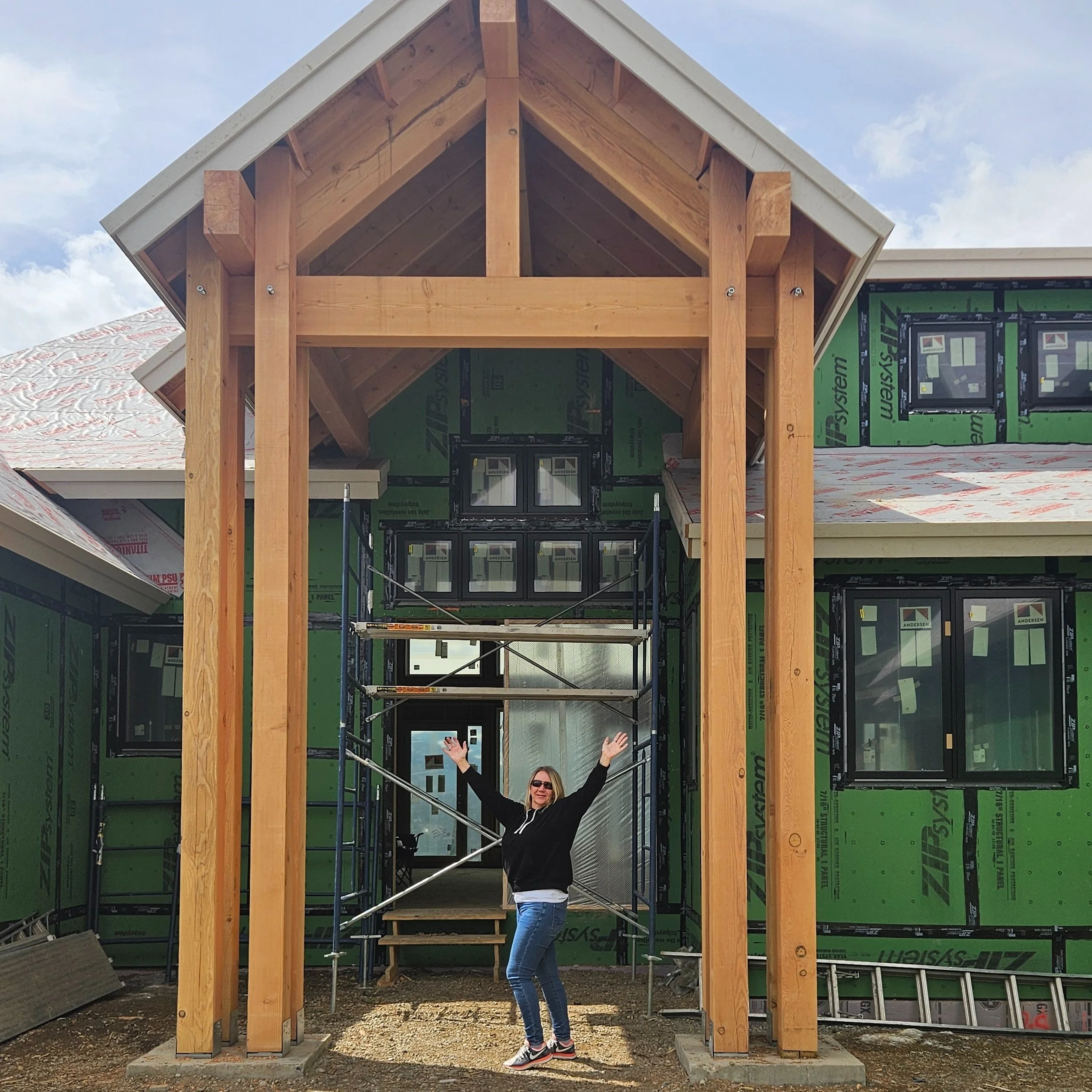 A woman standing in front of a partially constructed house, smiling with her arms raised, surrounded by scaffolding and wooden beams, with a house under construction in the background.