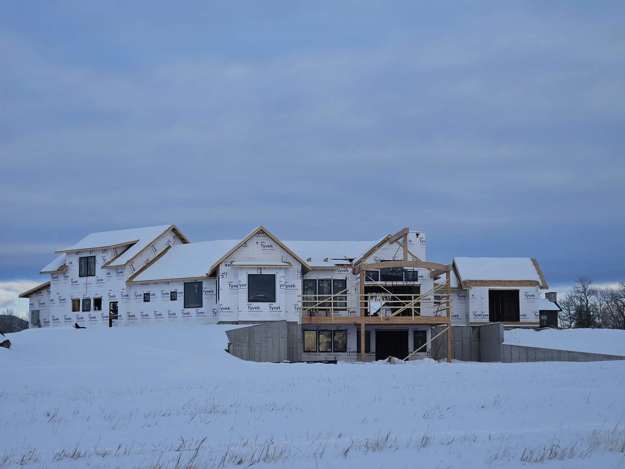 A house under construction in a snowy landscape, with wooden framing and partially installed siding.