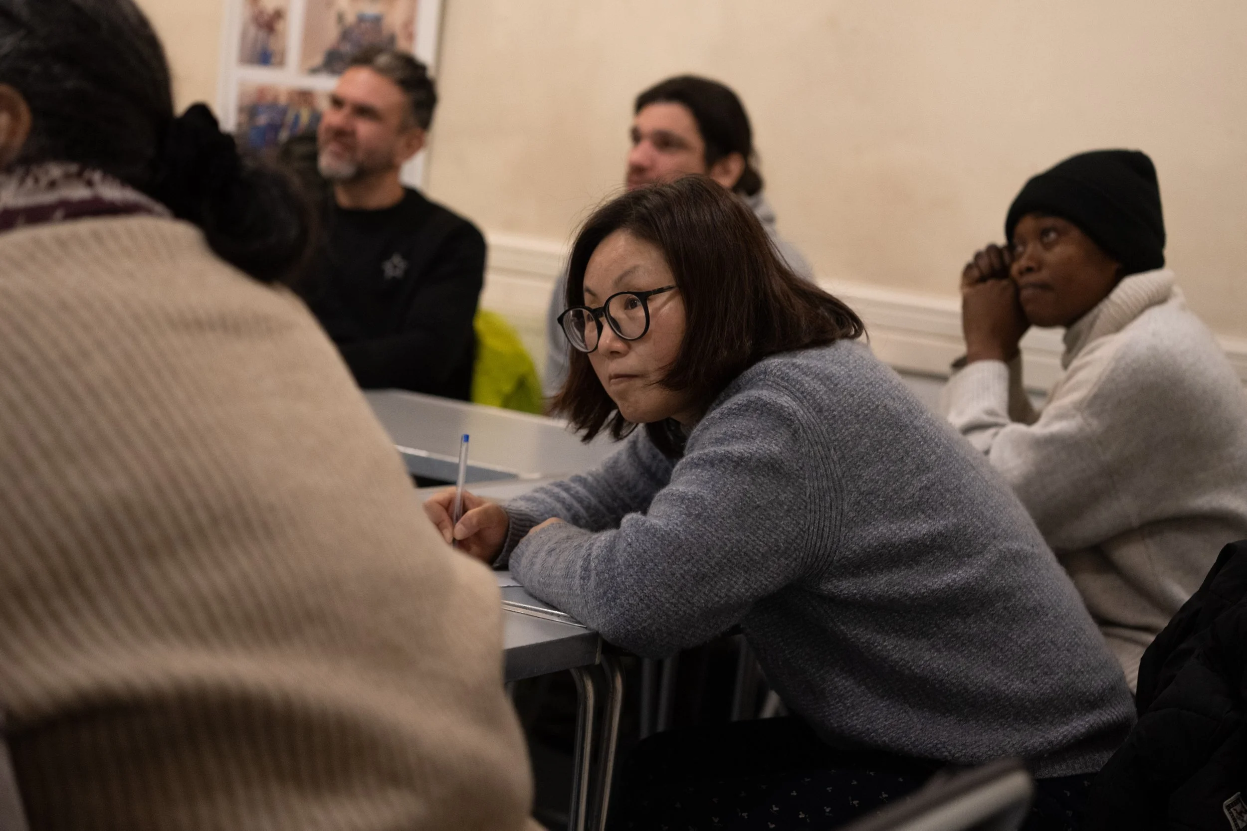 A woman with glasses taking notes, sitting at a table during a meeting or class. Other attendees are visible in the background.