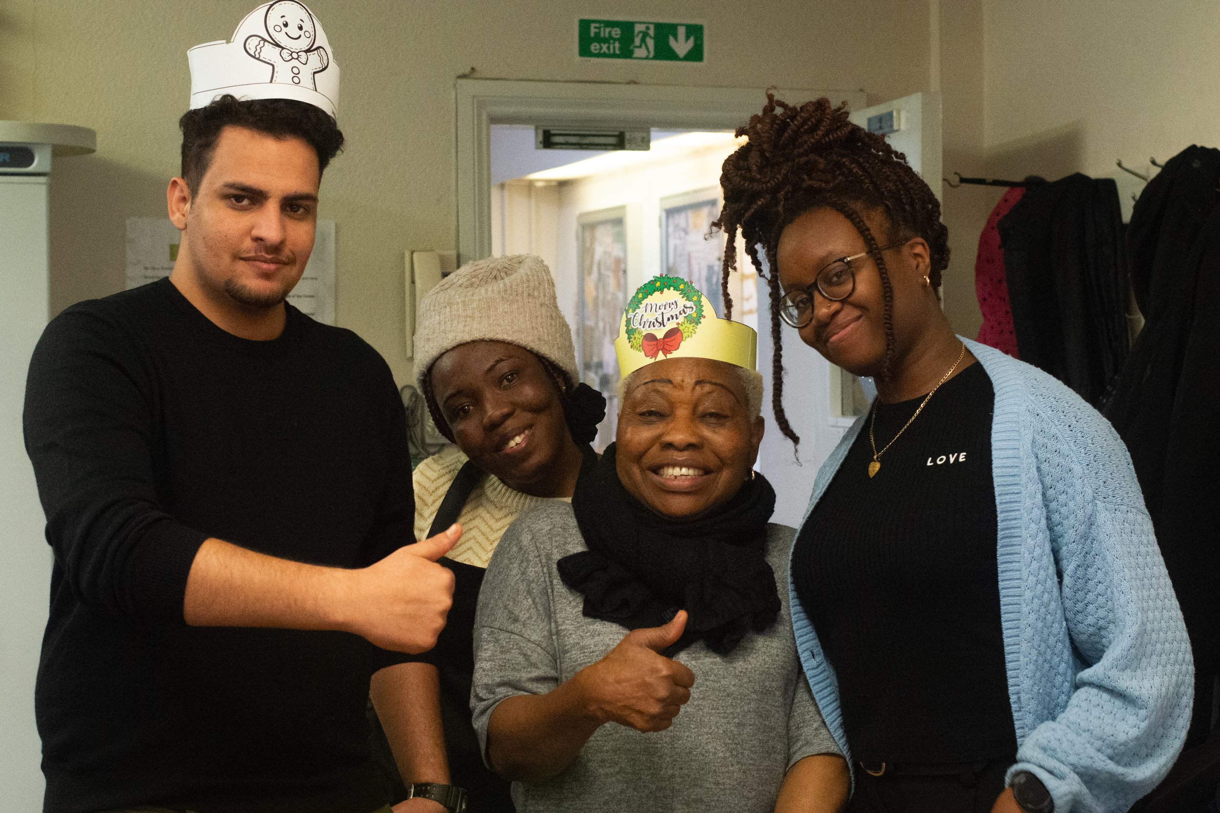 Group of four diverse women celebrating Christmas with a woman wearing a 'Merry Christmas' hat, all smiling and giving thumbs up in a cheerful indoor setting.