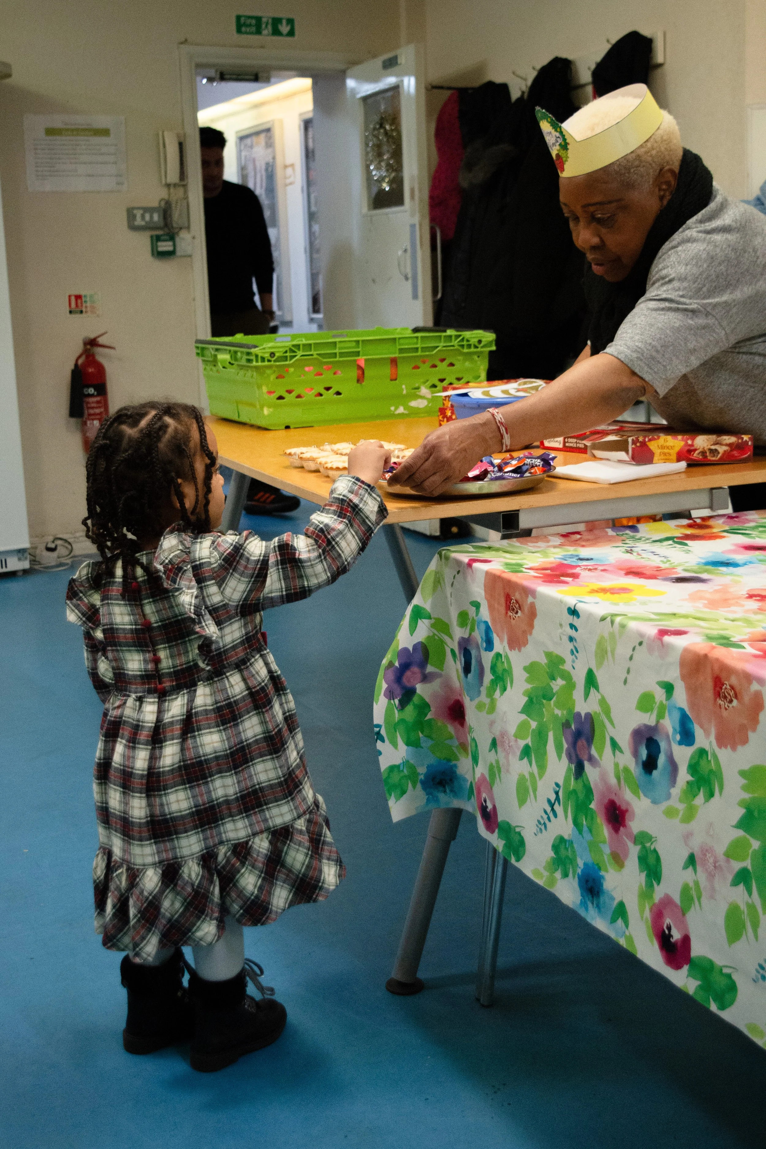 A young girl with braided hair, wearing a plaid dress and black boots, reaches out to receive a cookie from an older woman with short gray hair, wearing a paper party hat and a gray t-shirt. They are at a table with a floral tablecloth, and various b